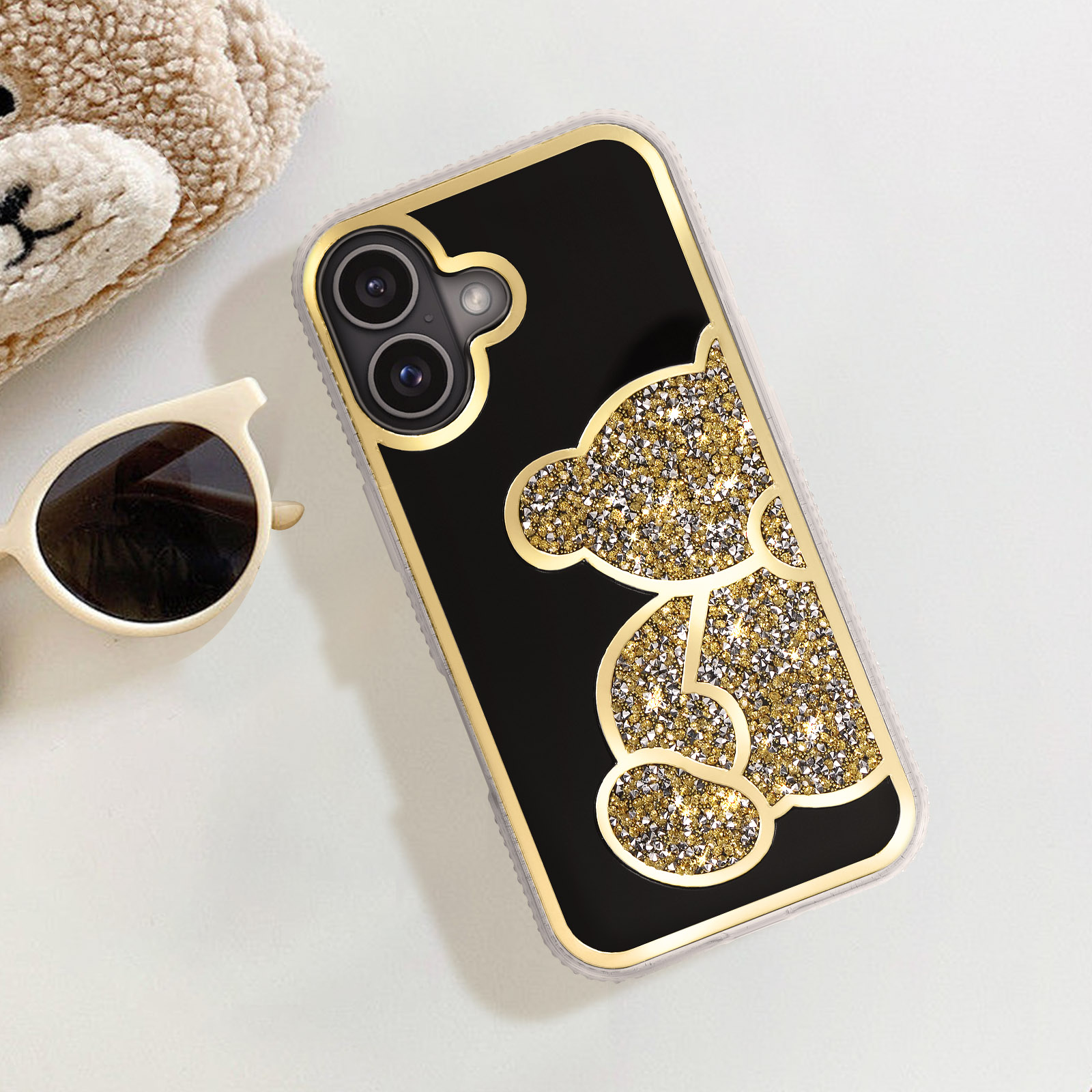 Eine Handyhülle mit goldglitzerndem Teddybären-Design. Sonnenbrille und Teddybär daneben.