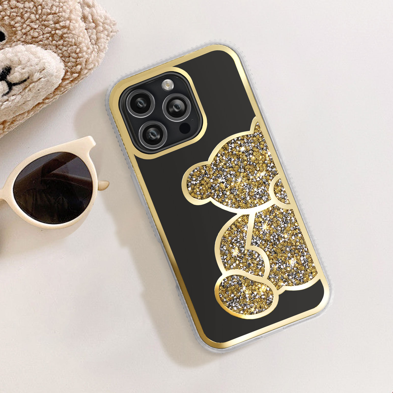 Eine Handyhülle mit goldenem Rahmen und glitzerndem Teddybären-Design, neben Sonnenbrille und Teddybär.