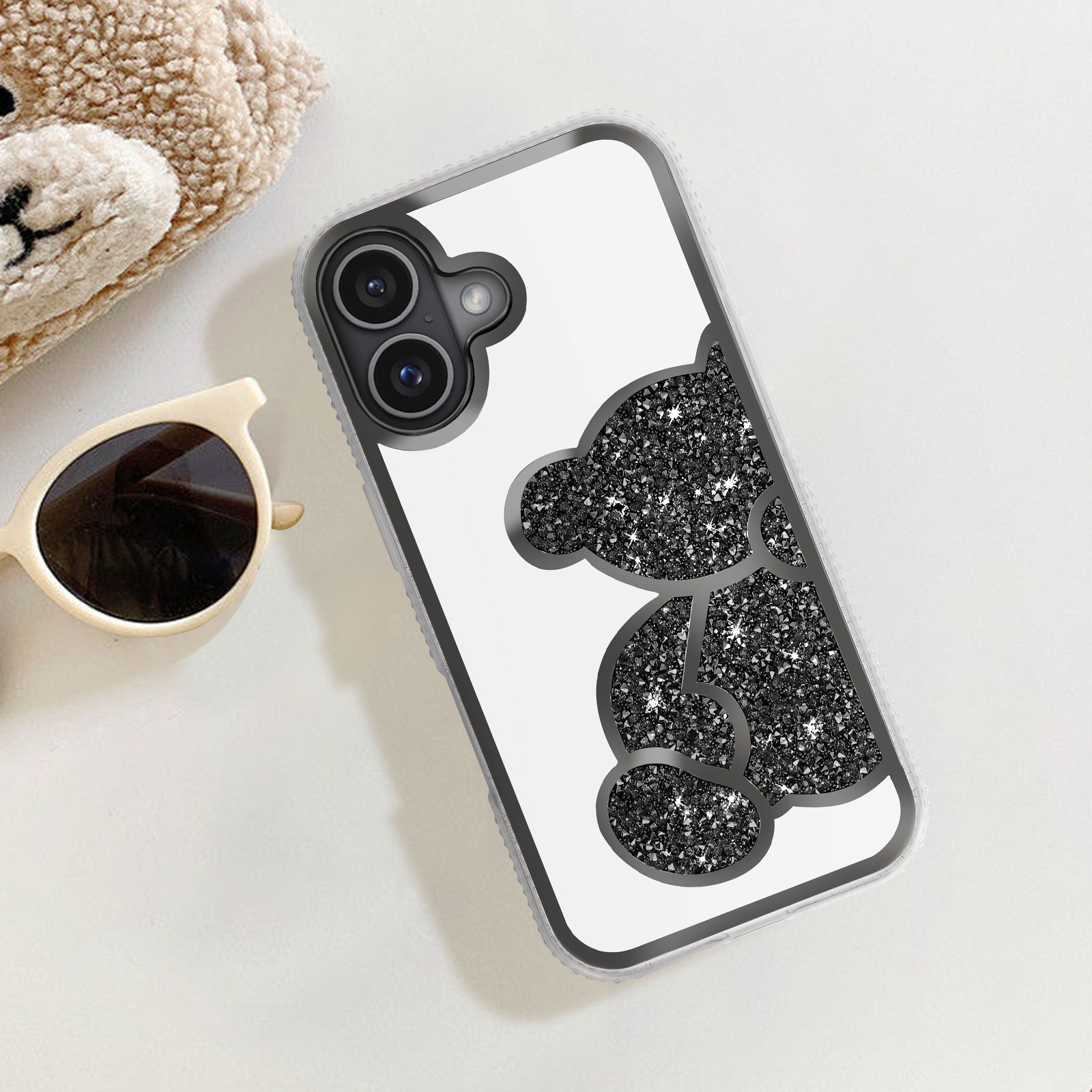 Custodia per telefono con un design a orsetto glitterato. Occhiali da sole e un orsacchiotto sono sulla superficie bianca. Lo sfondo è bianco.