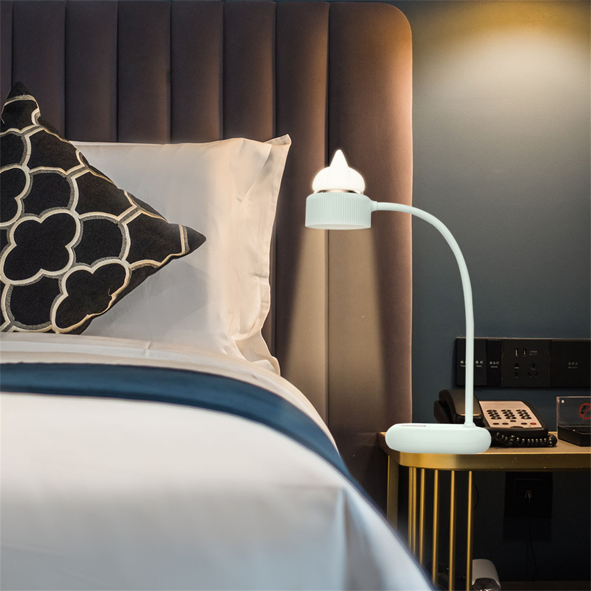 Eine weiße Schreibtischlampe mit gebogenem Arm steht auf einem kleinen Tisch. Ein Bett und ein Fenster sind im Hintergrund.
