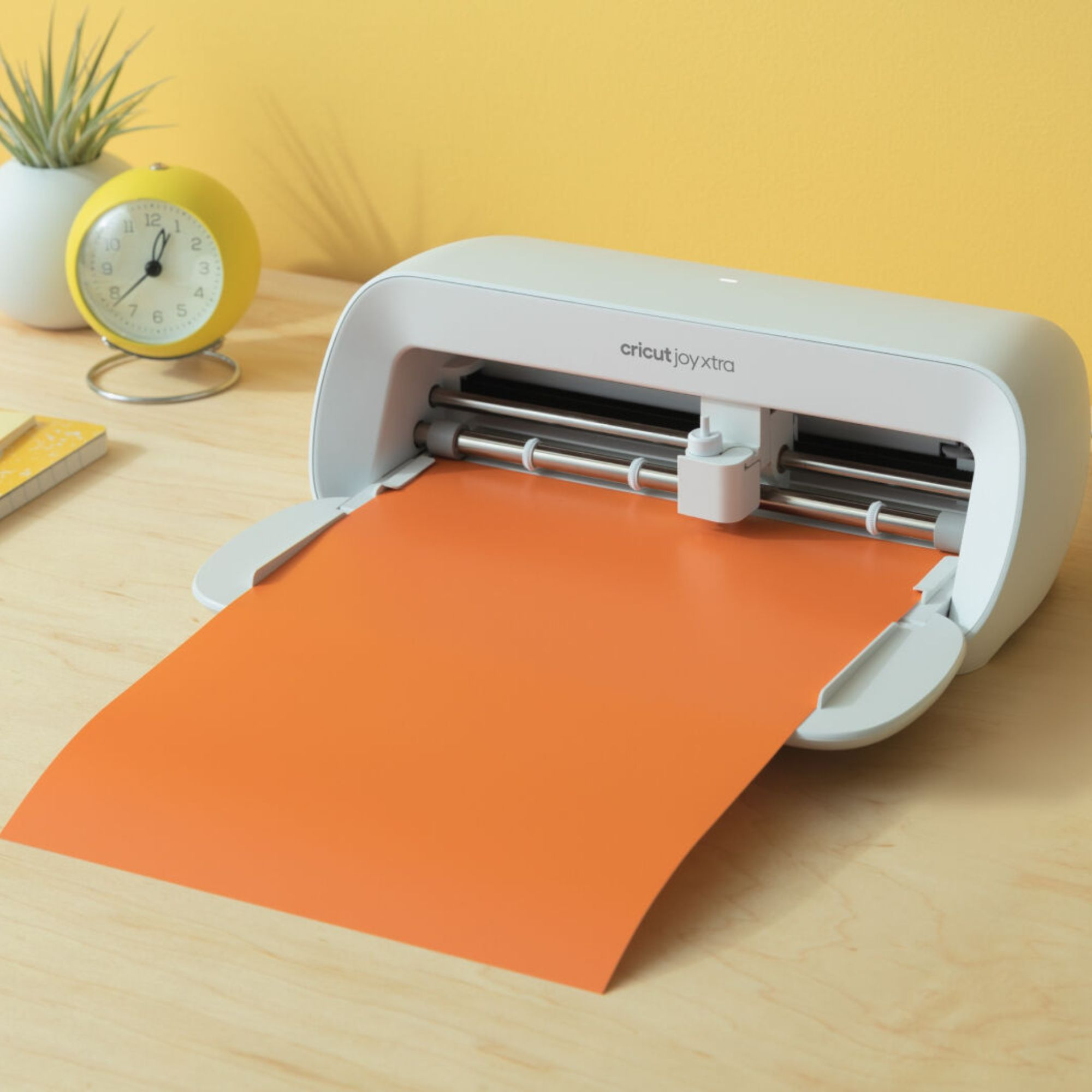 Máquina Cricut con papel naranja sobre una mesa de madera, junto a reloj amarillo y planta.