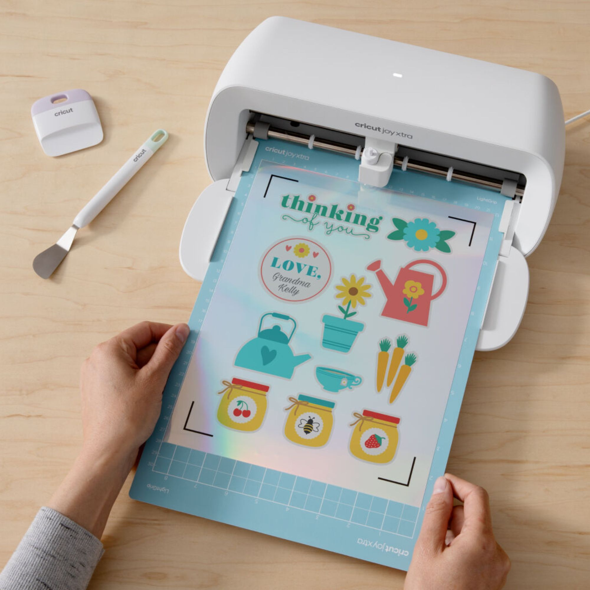 Persona sosteniendo una máquina Cricut con una hoja de pegatinas. Las pegatinas dicen 'thinking of you' y muestran imágenes.