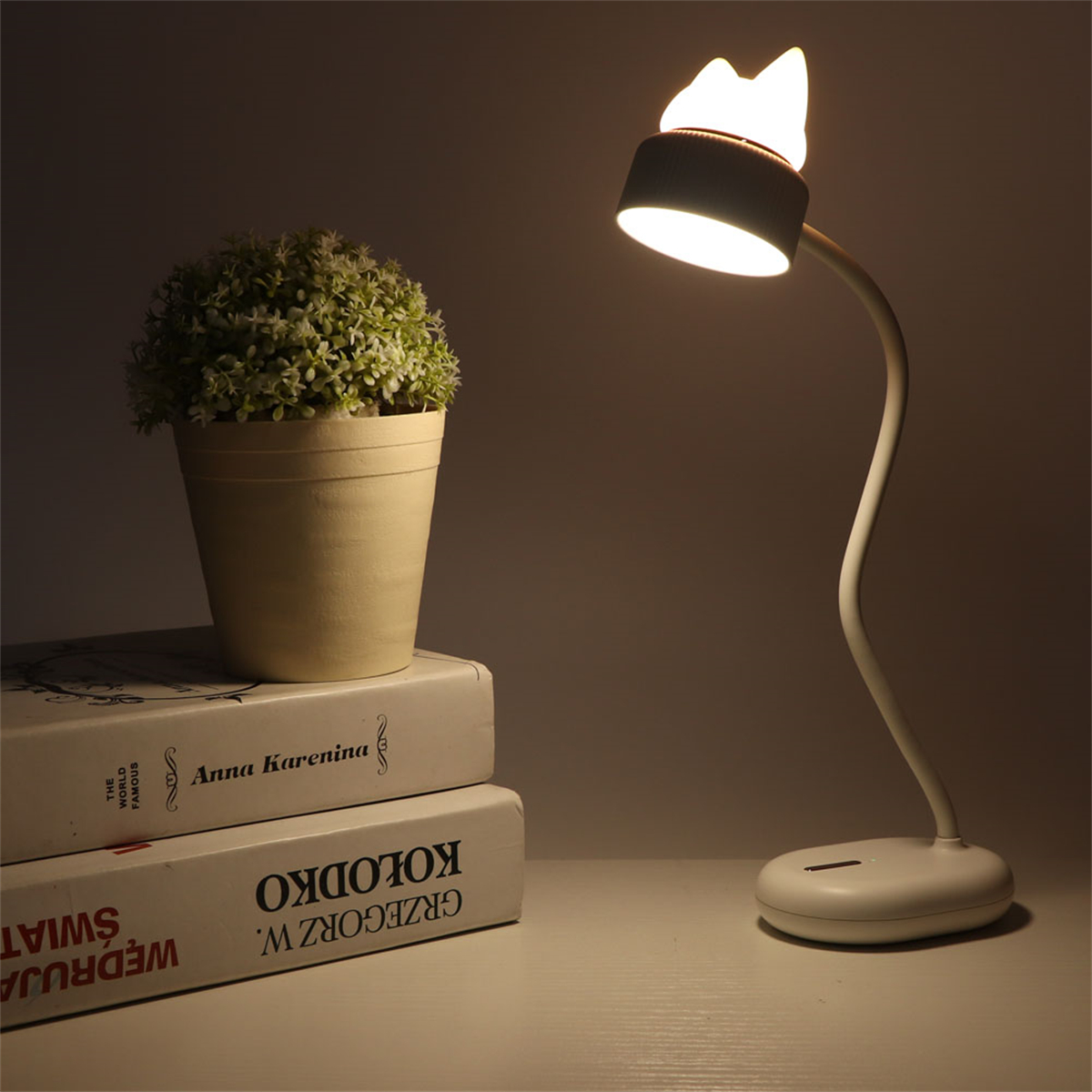 Katzenförmige Schreibtischlampe mit Buch und Pflanze auf weißer Oberfläche.