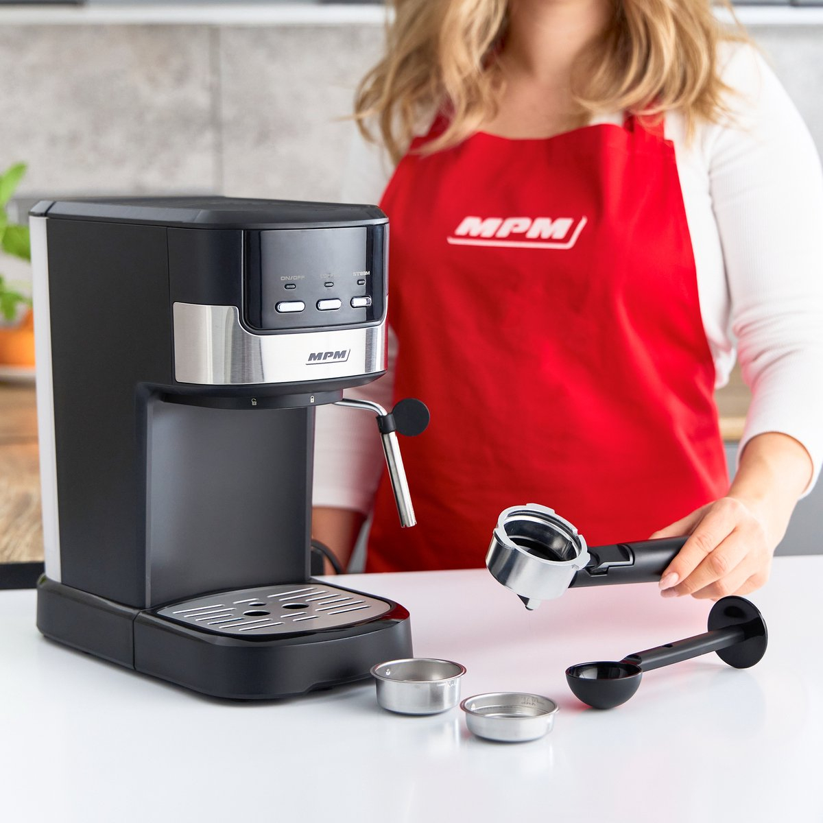 Macchina per caffè espresso. Donna con grembiule rosso, tiene il portafiltro. Accessori sul bancone.