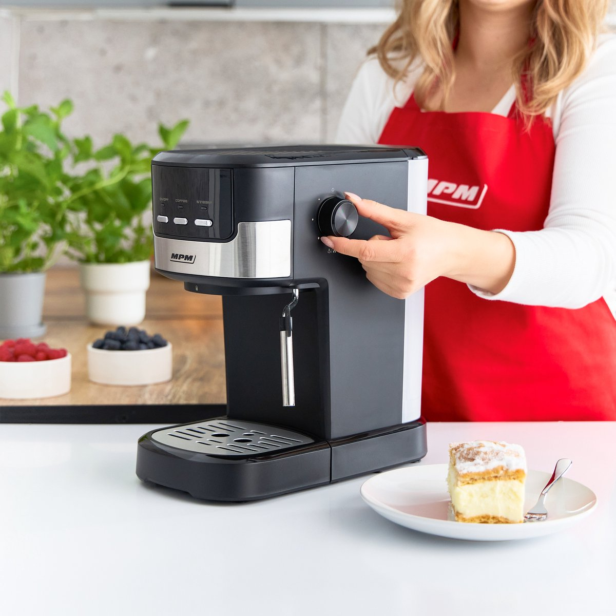 Donna che aziona una macchina da caffè nera e argento, con un piatto di torta e bacche su un tavolo.