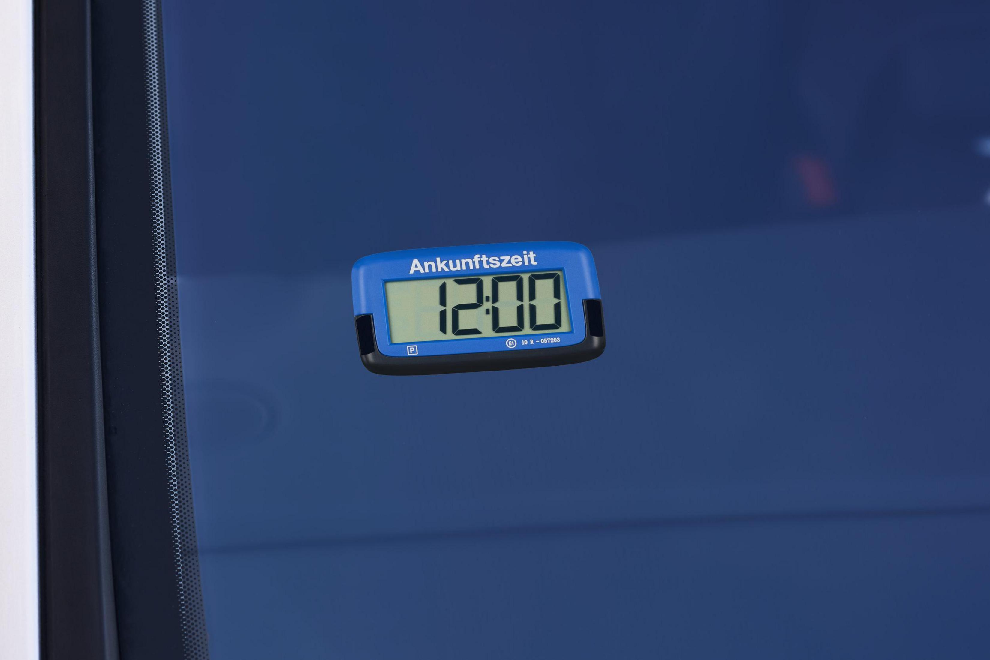 Ein blau-schwarzer digitaler Parktimer, der 12:00 anzeigt, in einem Autofenster. Der Text lautet 'Ankunftszeit'.