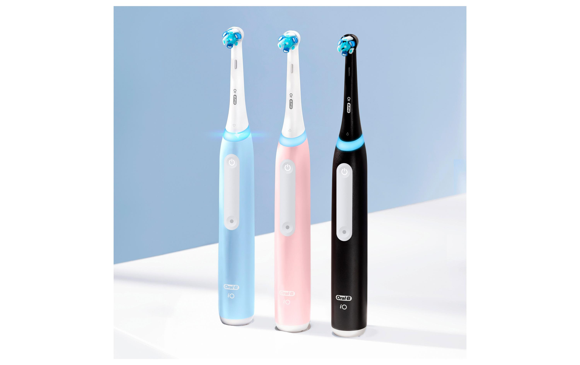 Trzy elektryczne szczoteczki do zębów, w kolorze niebieskim, różowym i czarnym, na białej powierzchni, z jasnoniebieskim tłem.