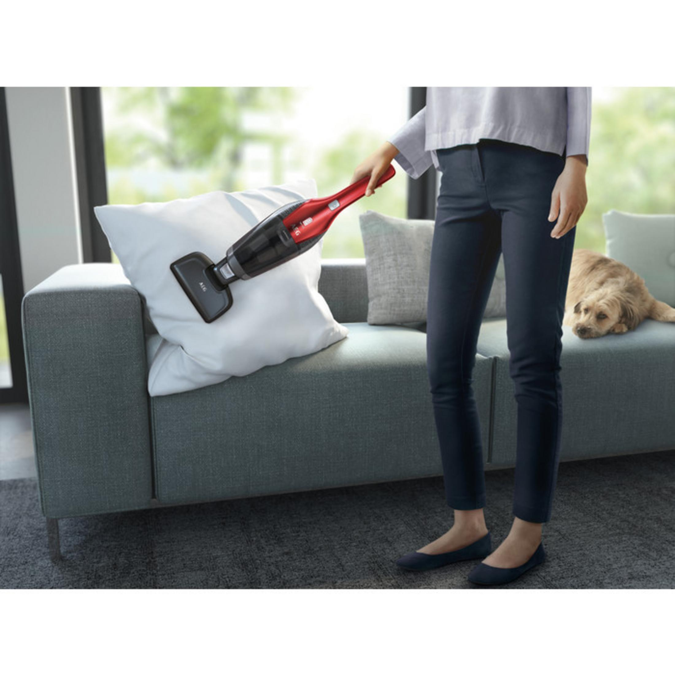 Una mujer aspira una almohada en un sofá gris con una aspiradora de mano roja.