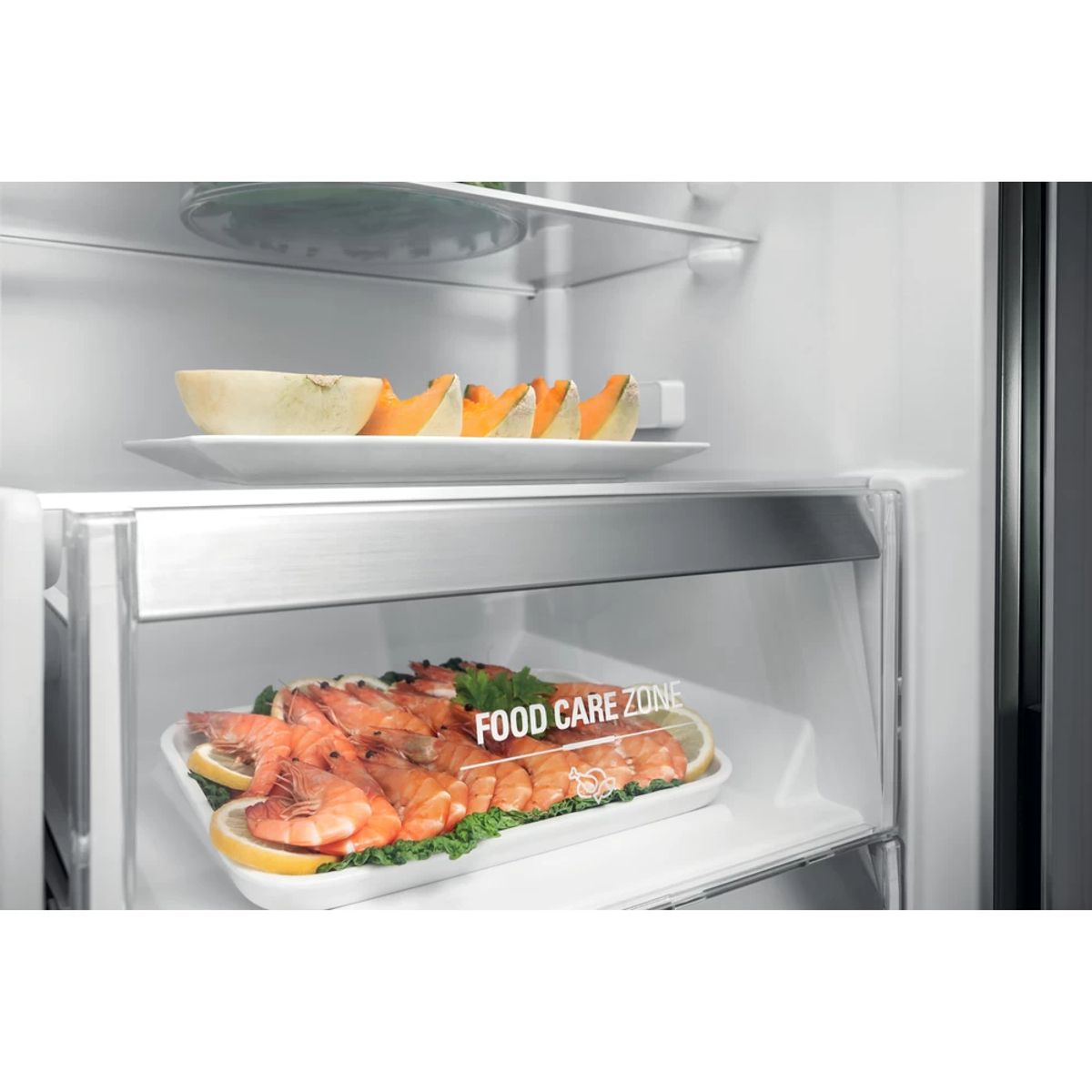 Dentro de una nevera, melón en rodajas en un plato, gambas y limones en bandeja. Etiqueta de zona de cuidado de alimentos.