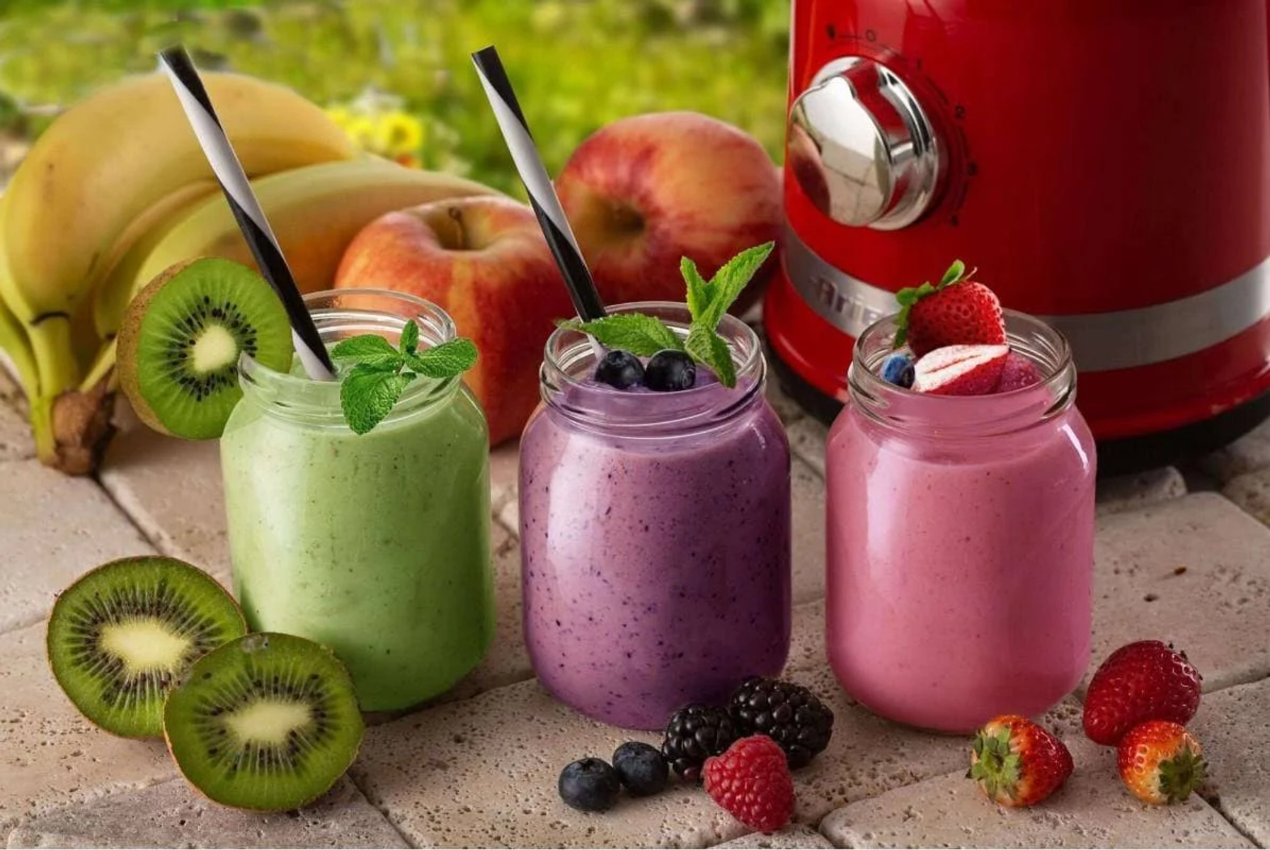 Drei Smoothies in Gläsern mit Strohhalmen, umgeben von Obst und einem roten Mixer.