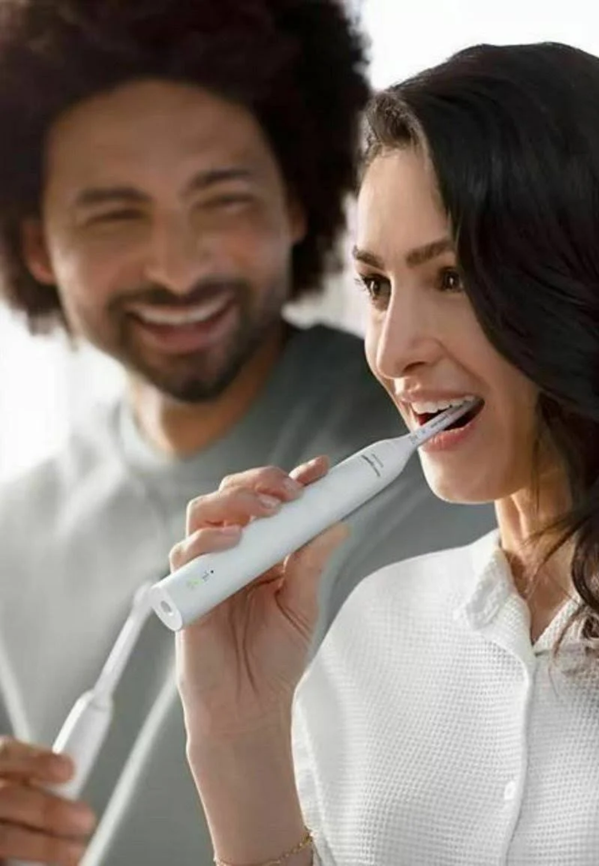 Kobieta używa białej elektrycznej szczoteczki. Mężczyzna w tle się uśmiecha. Biała łazienka.