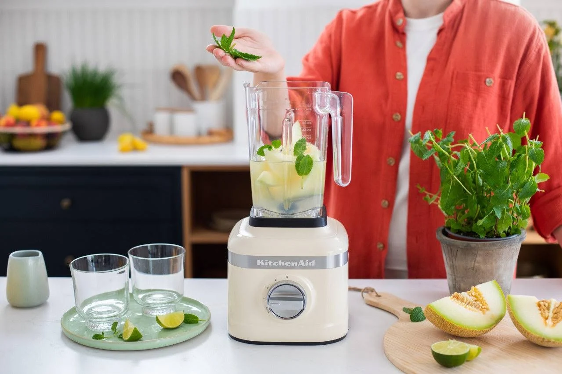 Osoba dodająca miętę do blendera z logo KitchenAid, z bliskimi składnikami i szklankami.