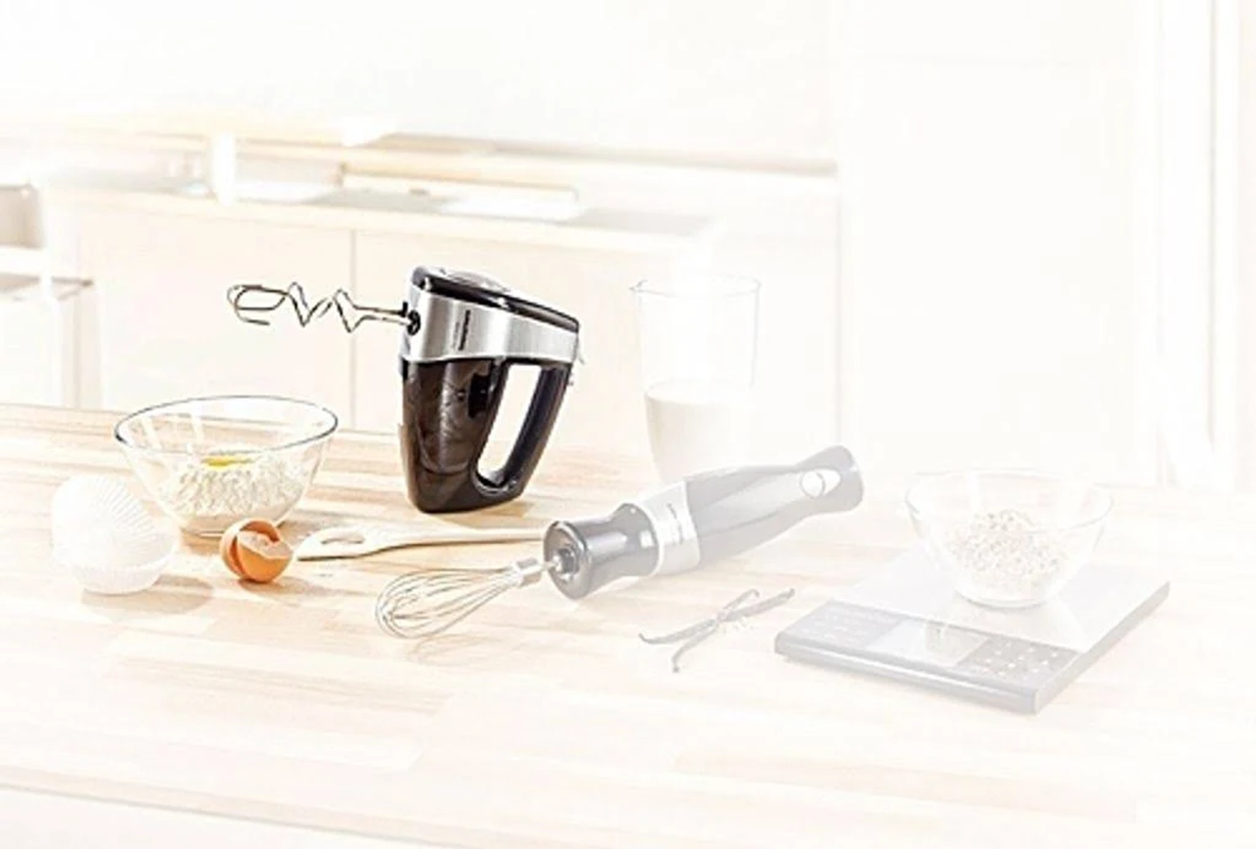 Un frullatore a mano con ingredienti come farina, uova e latte disposti su una superficie di legno.
