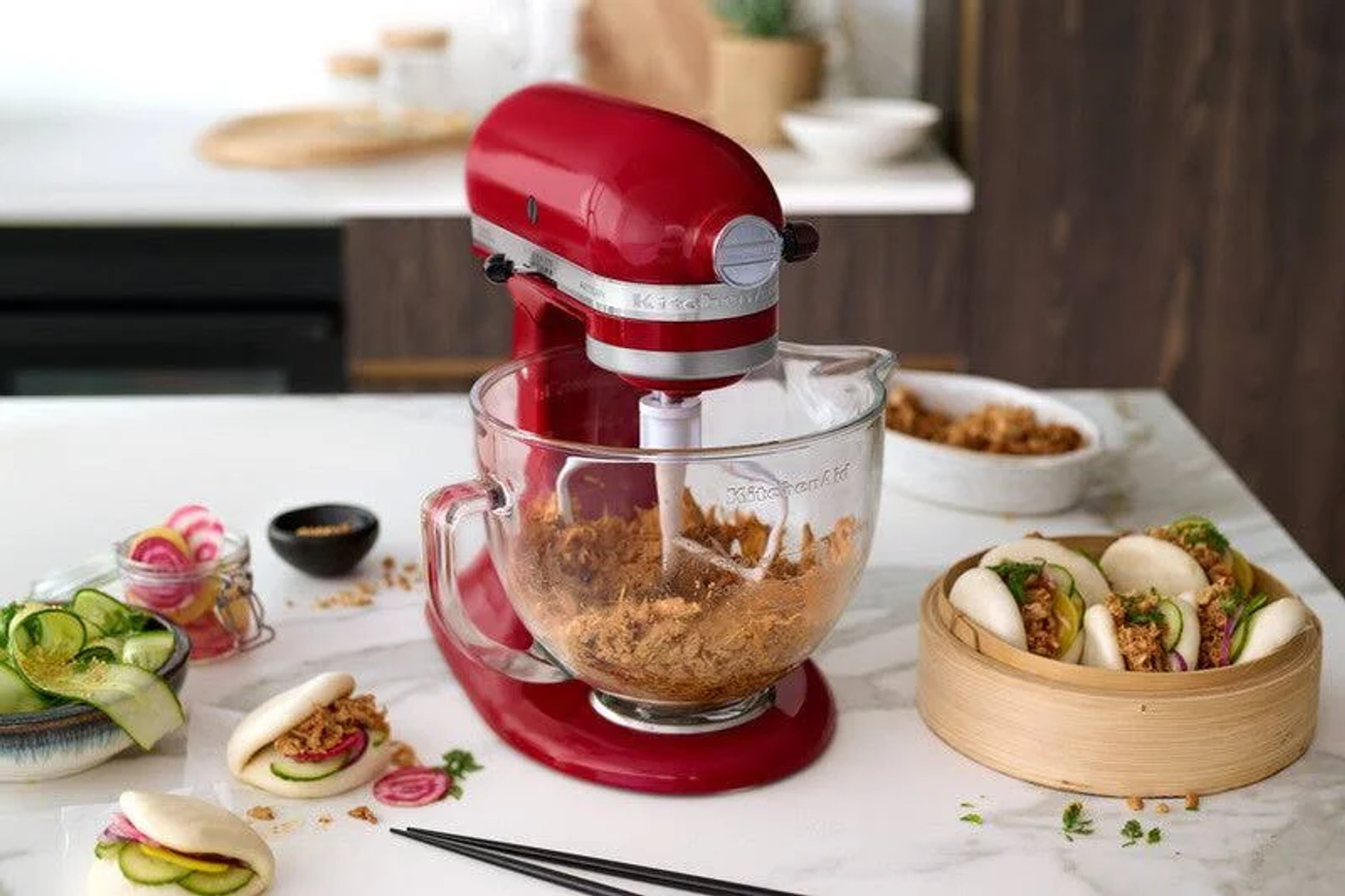 Roter KitchenAid-Mixer vermischt Zutaten in einer Glasschüssel. Gedämpfte Brötchen und andere Zutaten sind in der Nähe.