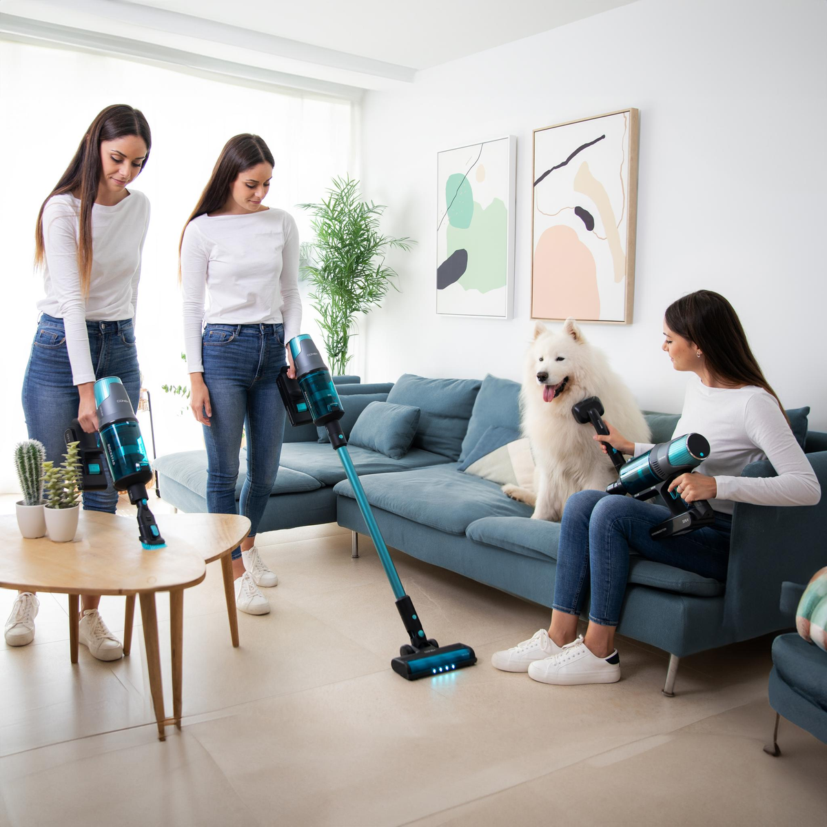 Tre donne e un cane in un soggiorno. Tengono in mano degli aspirapolvere. Divano e tavolo.