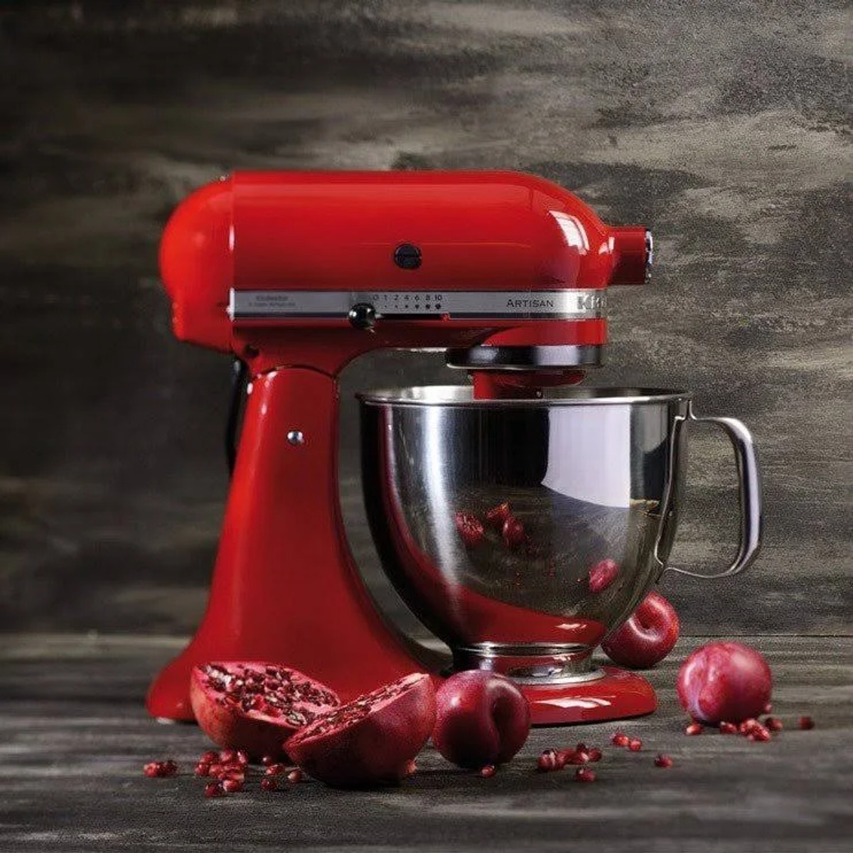 Kırmızı KitchenAid mikser, paslanmaz çelik kase, nar ve erik ahşap yüzey üzerinde.