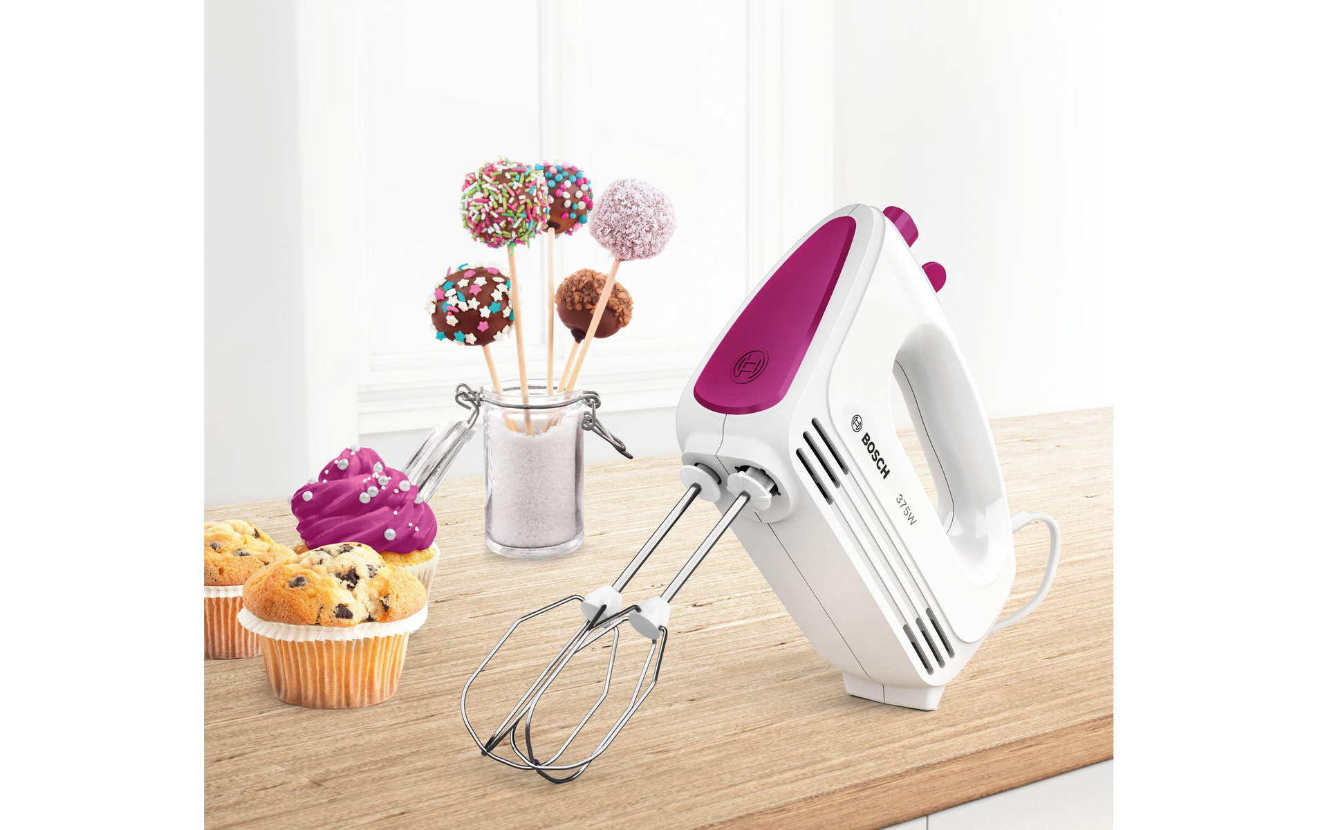 Ein weißer Handmixer mit rosa Oberteil, Cupcakes und Cake Pops auf einer Holzoberfläche und ein Glas Zucker.