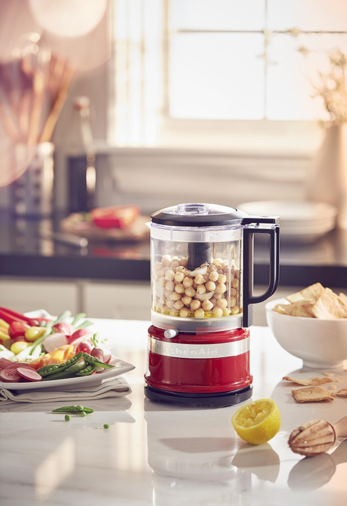 Kırmızı bir KitchenAid mini mutfak robotu, nohutla dolu, tezgahta başka malzemelerle.
