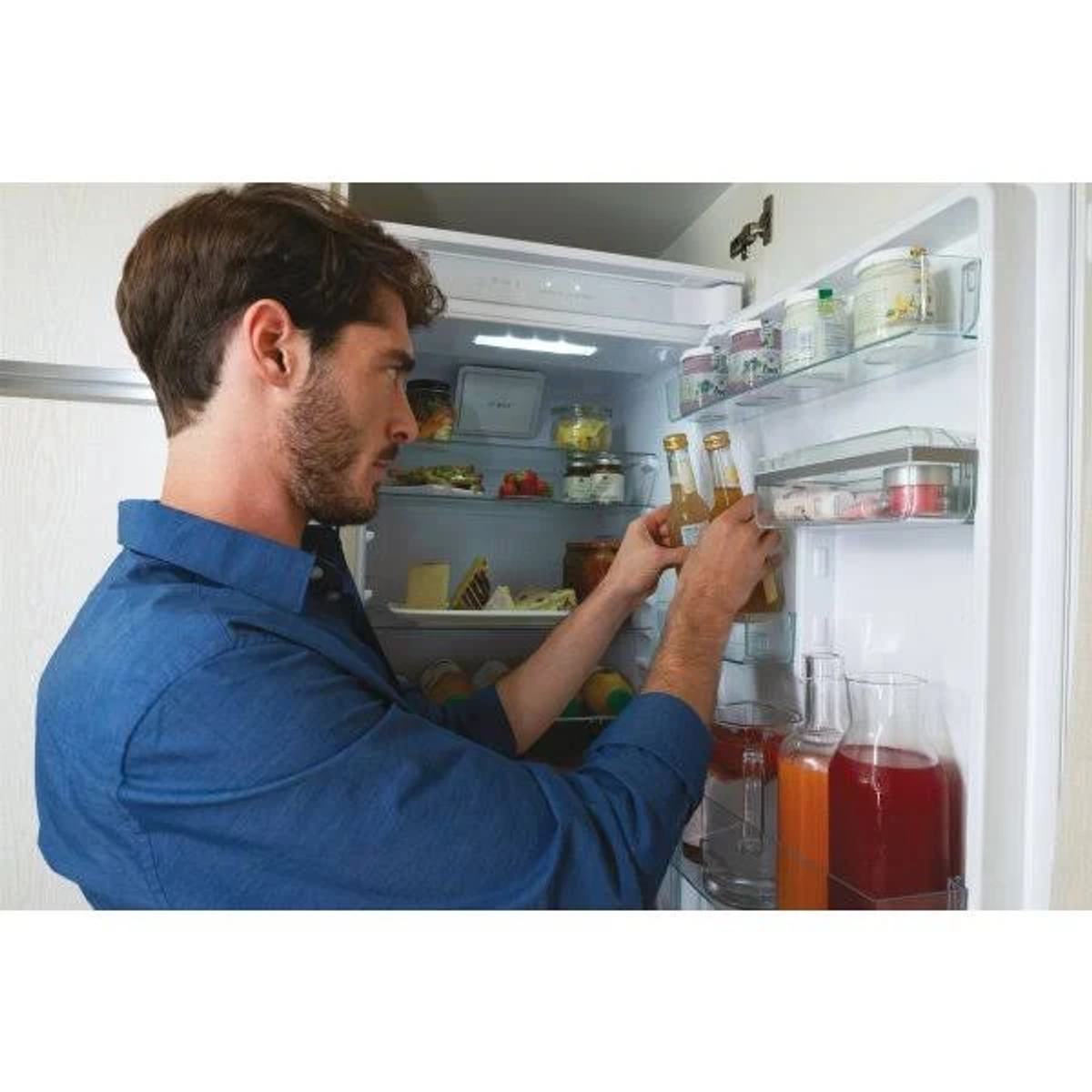 Mężczyzna w niebieskiej koszuli wyjmuje butelki z lodówki pełnej jedzenia i napojów.