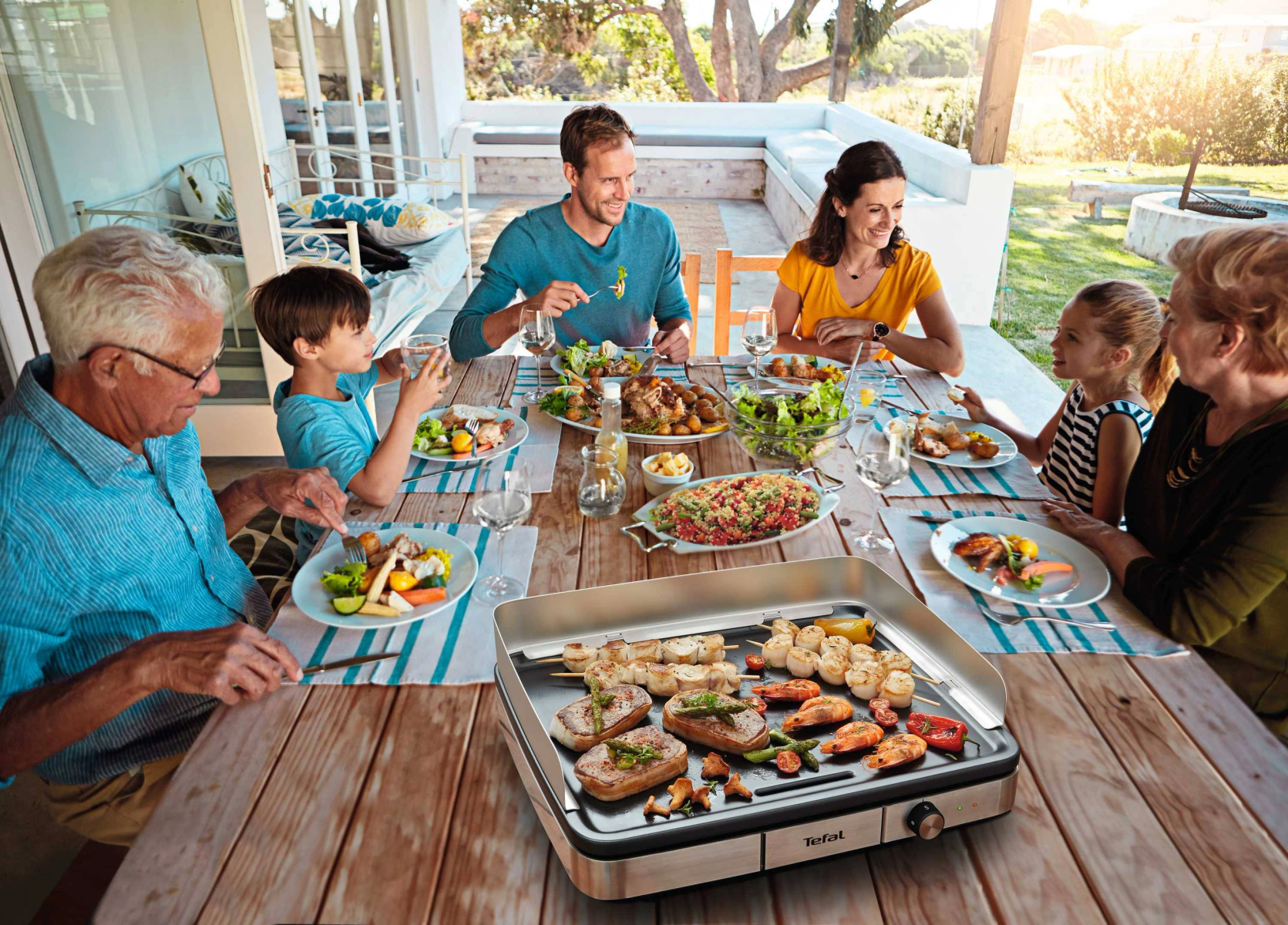Rodzina spożywająca posiłek na świeżym powietrzu, z grillem na drewnianym stole.