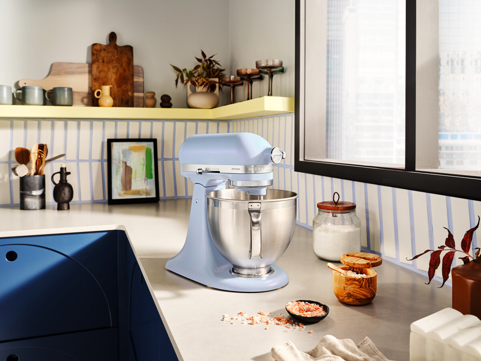 Impastatrice KitchenAid azzurra su un piano di lavoro, con oggetti da cucina sullo sfondo.