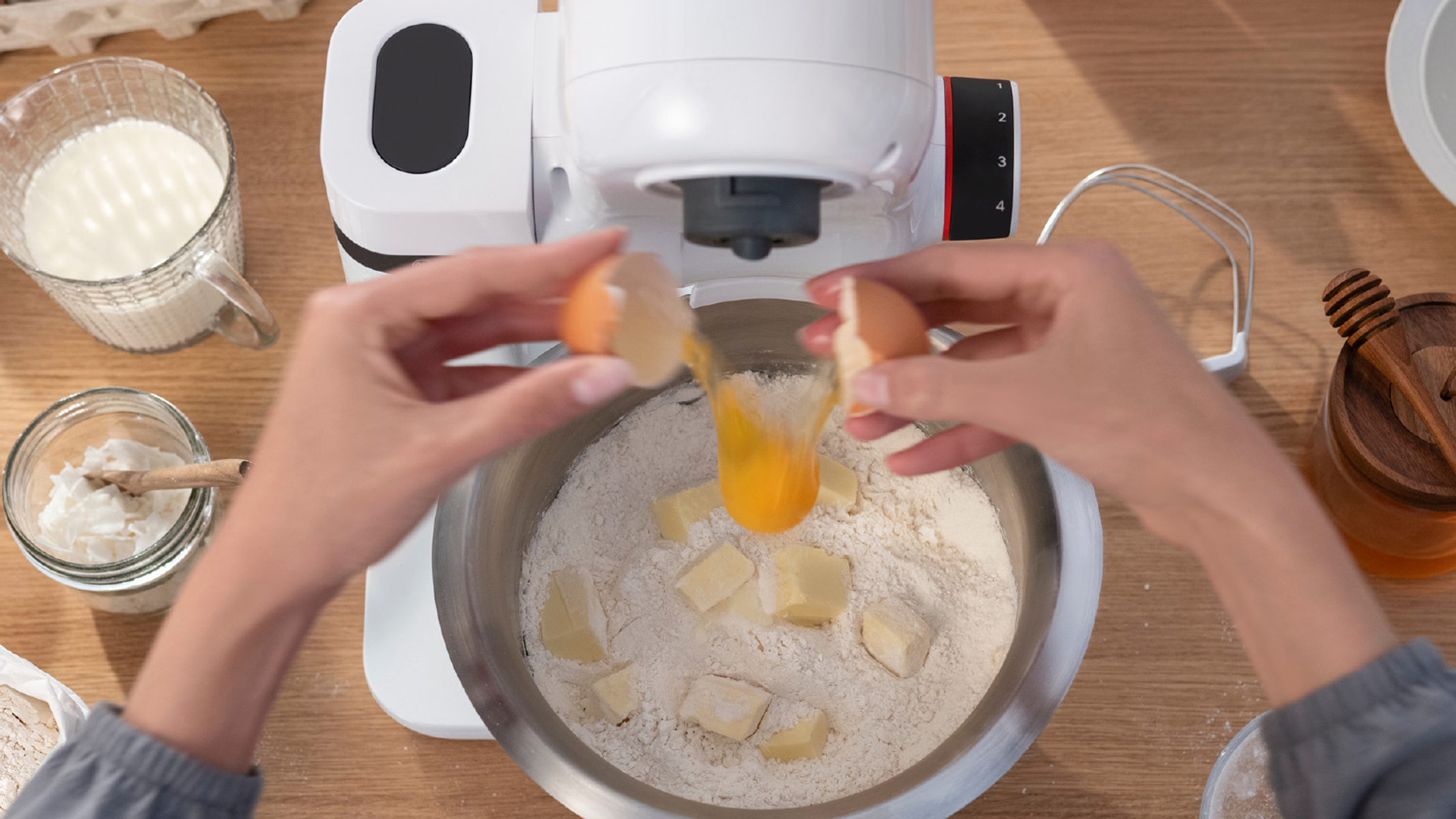 Manos rompiendo un huevo en un cuenco plateado con harina, cubos de mantequilla y otros ingredientes.