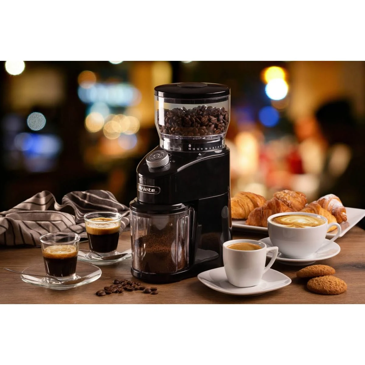 Kaffeemühle mit Kaffeebohnen, gemahlenem Kaffee, Espresso und Croissants auf einem Tisch.