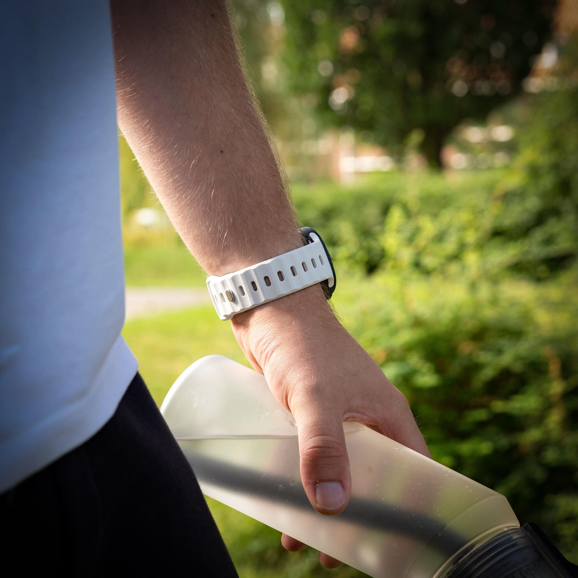 Ein Arm mit einer weißen Uhr und einer Wasserflasche vor grünem Buschwerk.
