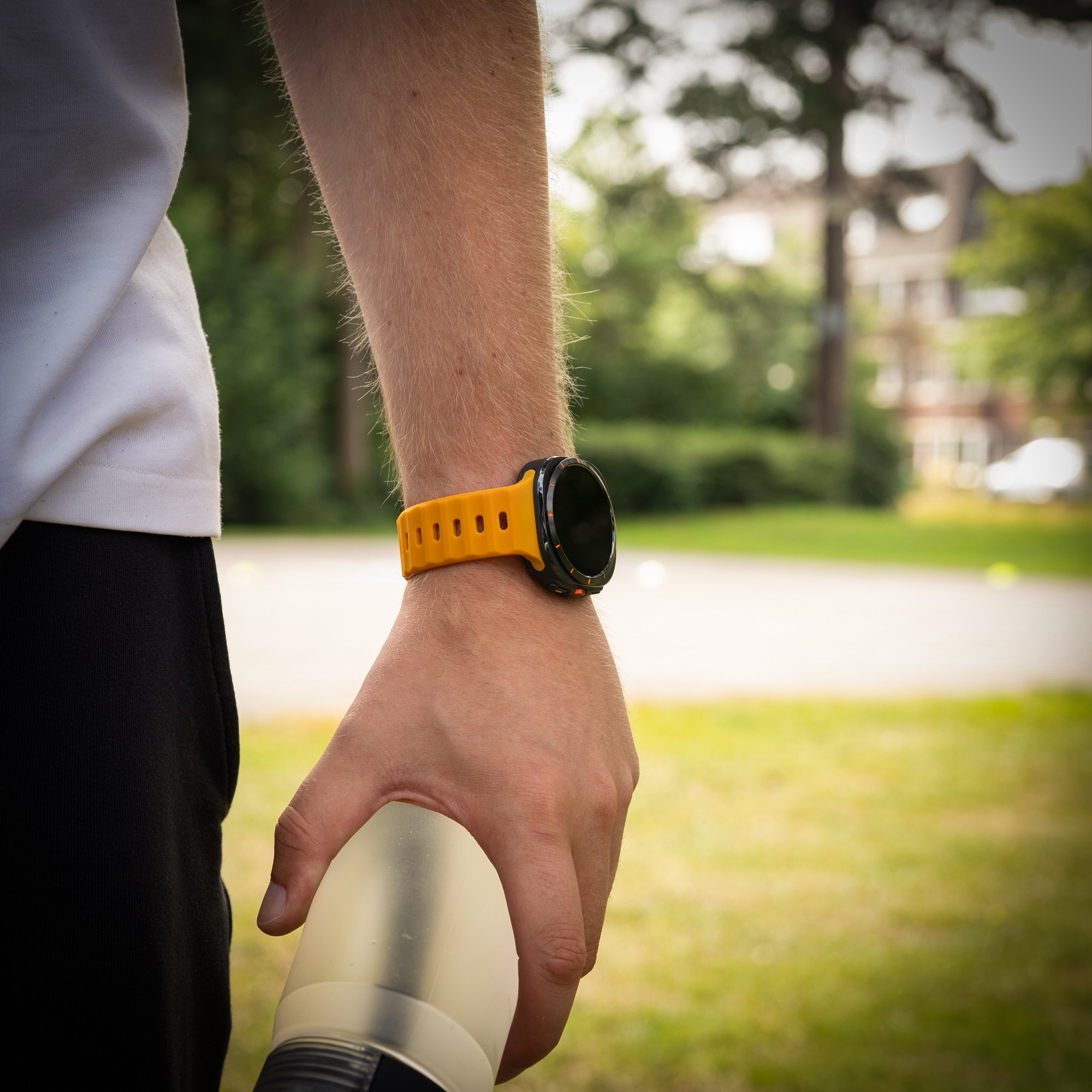 Eine Person trägt eine orangefarbene Smartwatch an ihrem linken Handgelenk und hält eine Wasserflasche in einem grasbewachsenen Feld.