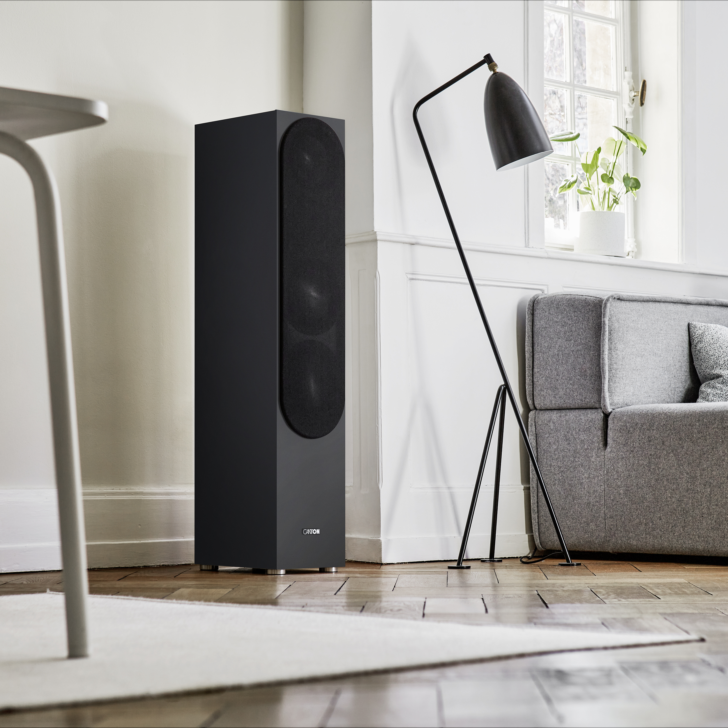 Schwarzer Lautsprecher neben einer Stehlampe und einem grauen Sofa, Holzboden und weiße Wand.