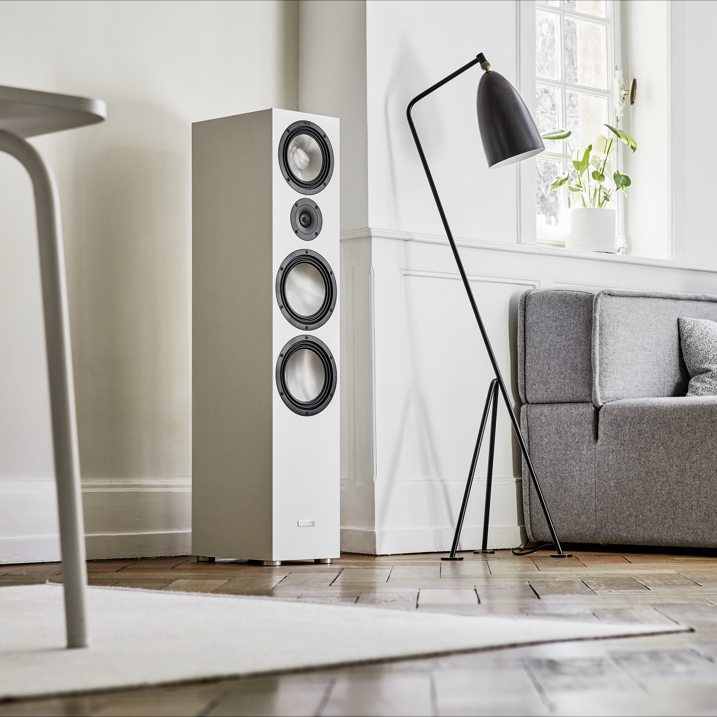 Weißer hoher Lautsprecher neben schwarzer Stehlampe und grauem Sofa, auf Holzboden.