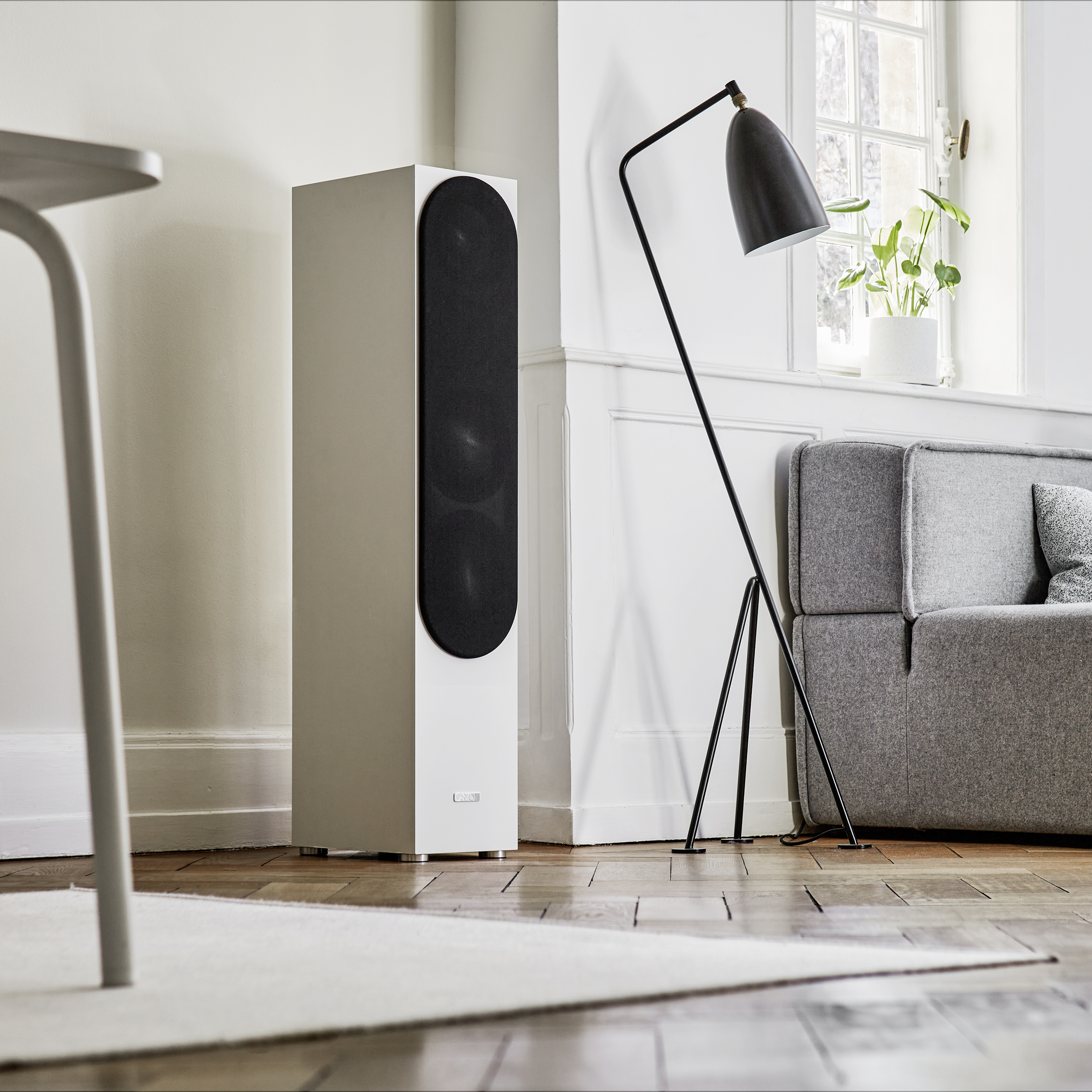 Ein weißer Lautsprecher auf einem Holzboden, eine schwarze Lampe, ein graues Sofa, ein Fenster und ein Teppich.