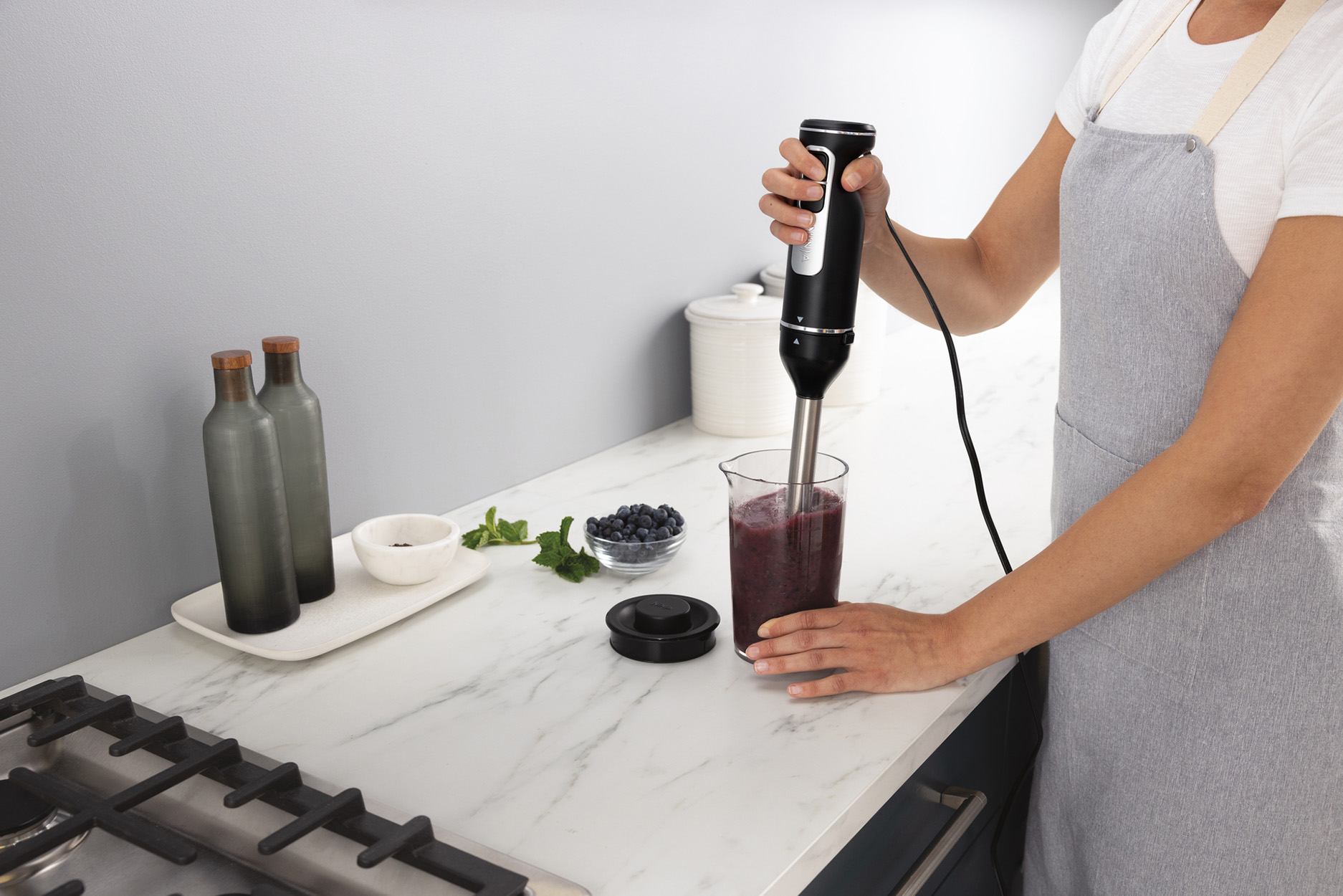 Eine Person, die einen Smoothie in einem Glas mit einem Handmixer mixt. Beeren und ein Glas sind zu sehen.