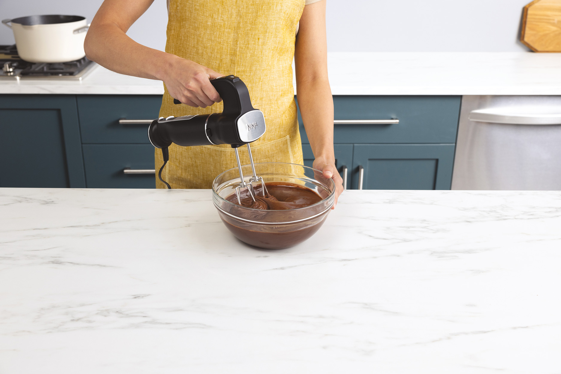 Eine Person in einer gelben Schürze verwendet einen schwarzen Handmixer, um Zutaten in einer Glasschüssel zu mischen.