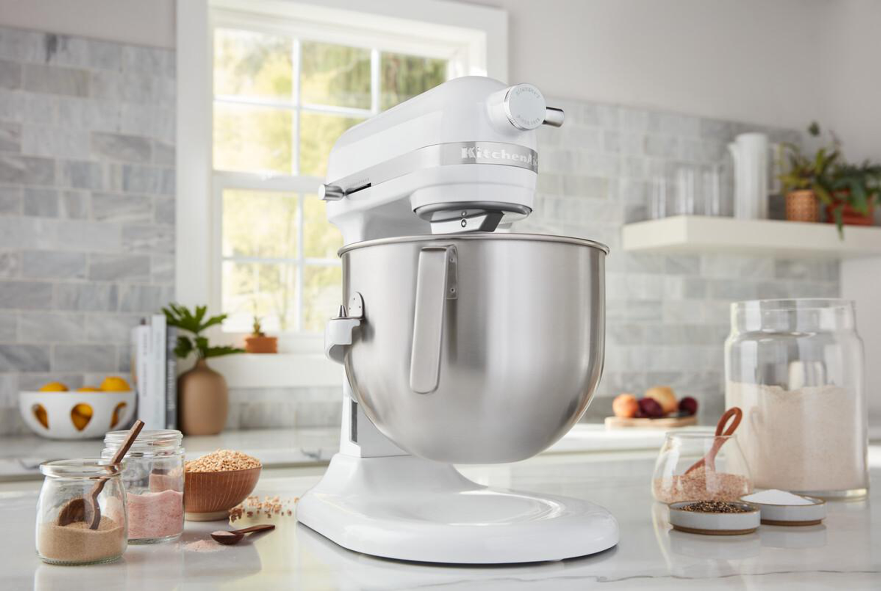 Weißer KitchenAid Mixer auf Marmortisch. Gläser mit Zutaten und Utensilien im Hintergrund.