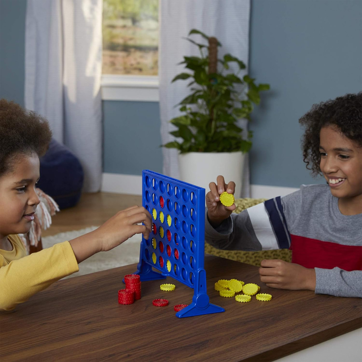 Zwei Kinder spielen Connect 4 auf einem braunen Tisch, lächelnd.
