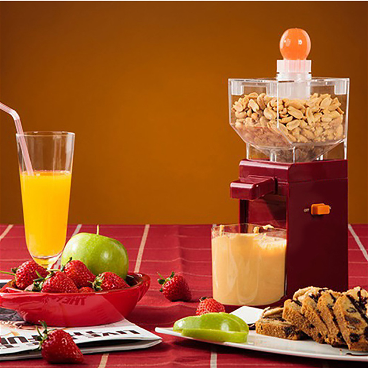 Eine rote Erdnussbuttermaschine mit Erdnüssen darin. Ein Glas Orangensaft und Erdbeeren sind davor.