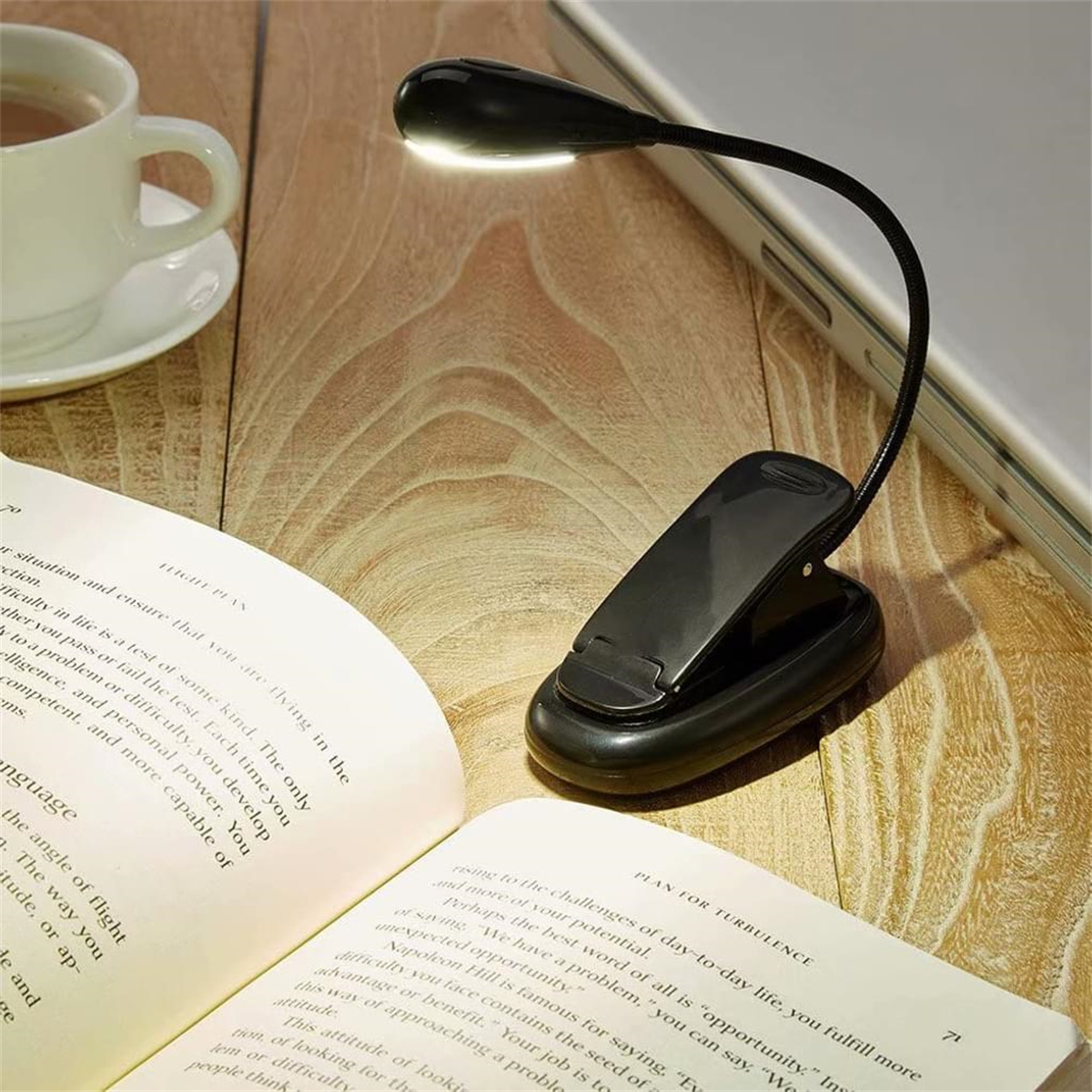 Eine schwarze Leselampe an einem Buch befestigt. Eine weiße Kaffeetasse und ein Laptop sind im Hintergrund.