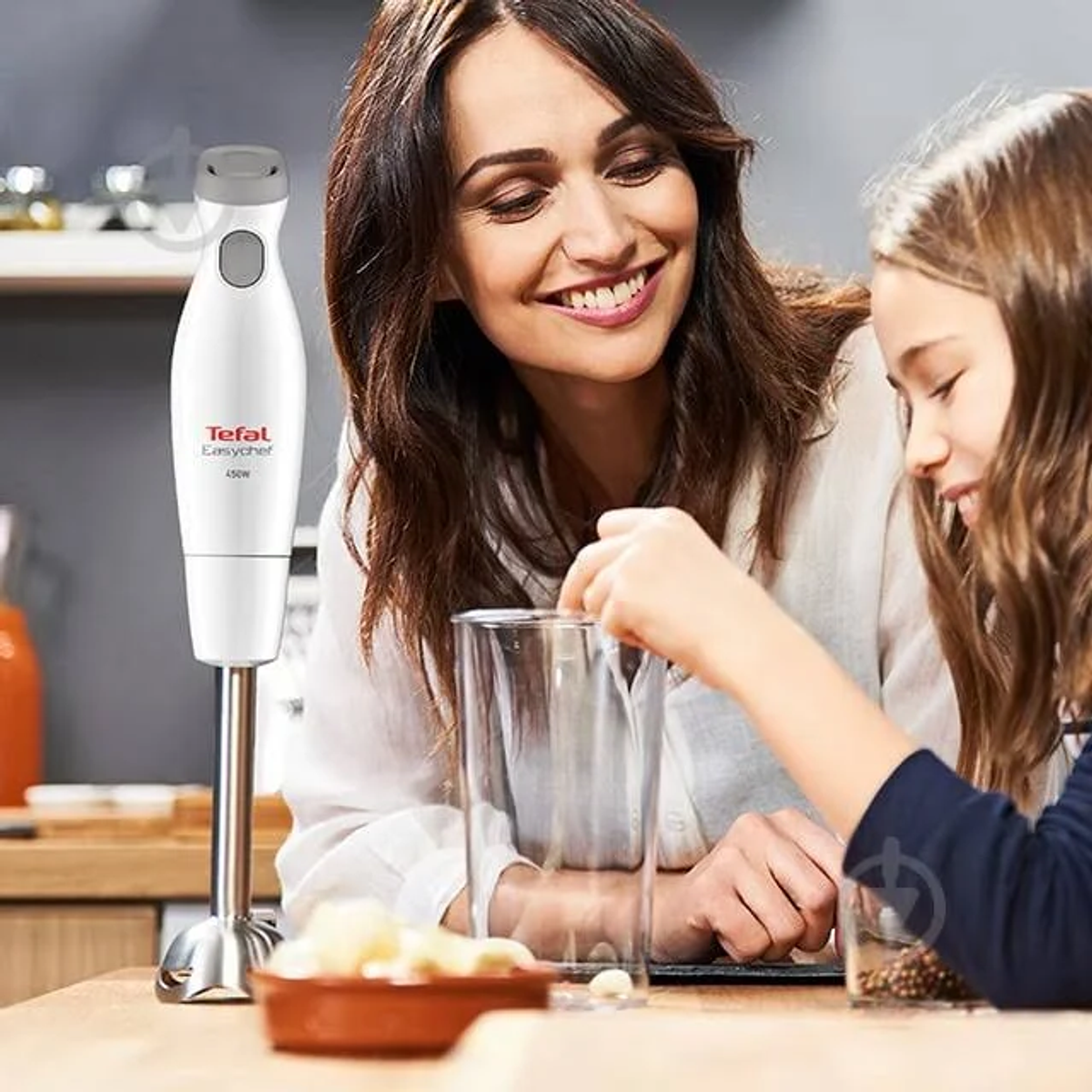 Vrouw en meisje gebruiken een Tefal staafmixer. Een glas en een kom staan op tafel.