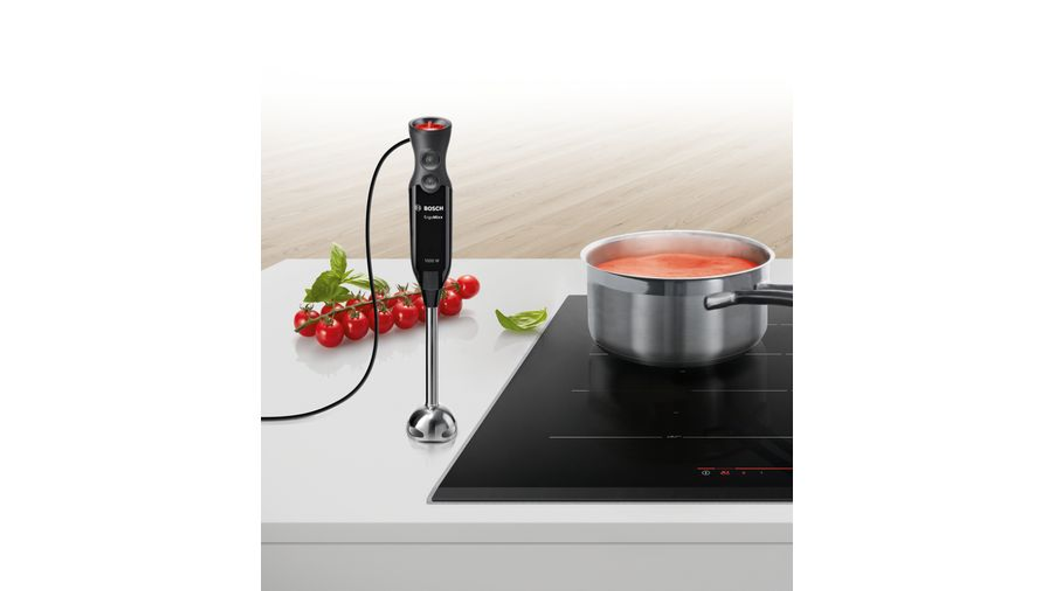 Czarny blender ręczny na białej powierzchni, z garnkiem zupy pomidorowej i pomidorami.