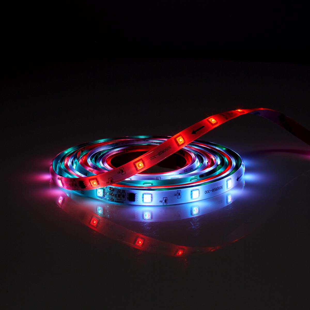 Eine aufgerollte LED-Leiste mit roten, blauen und grünen Lichtern auf reflektierender Fläche.