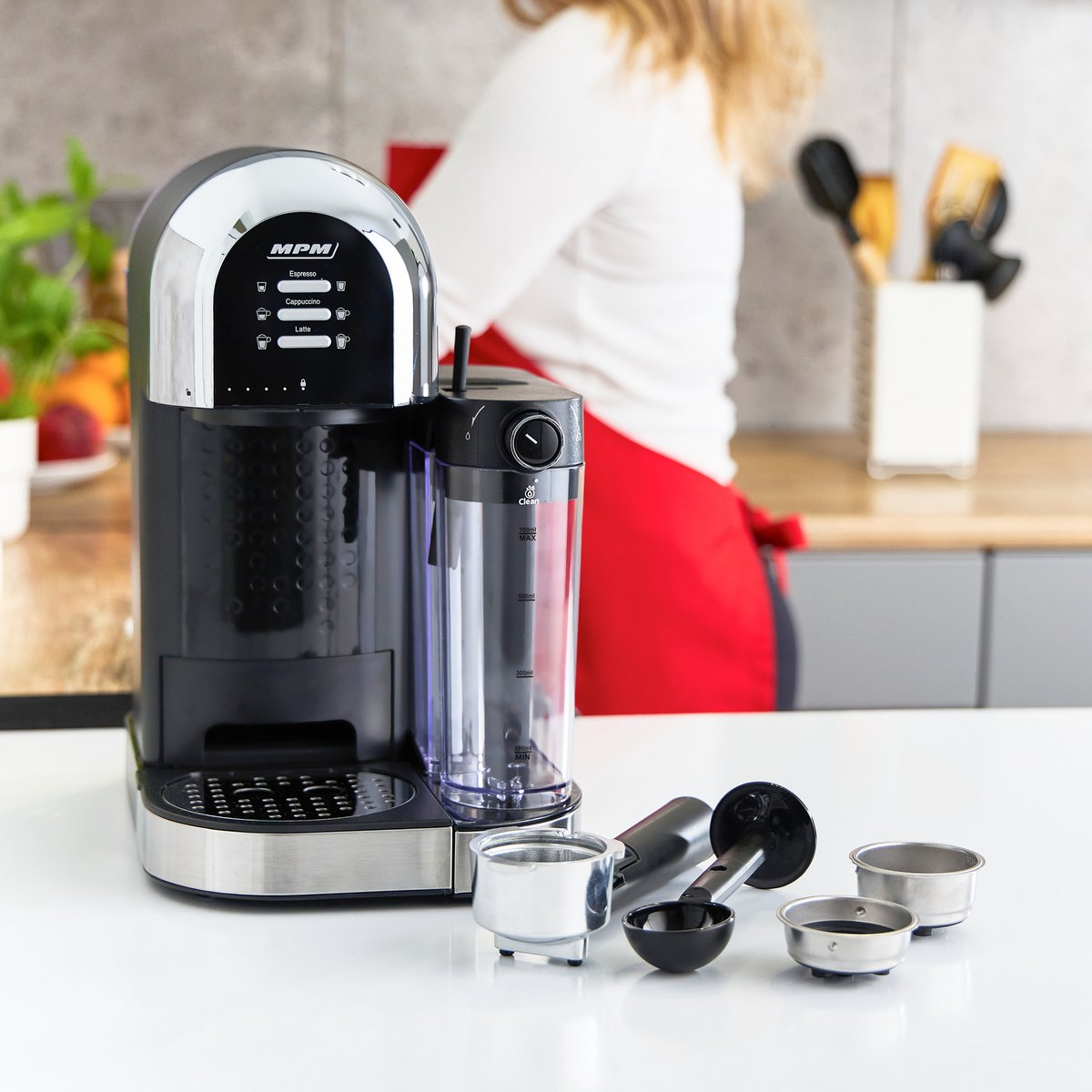 Schwarz-silberne Kaffeemaschine. Zubehör auf weißer Theke. Frau im Hintergrund.