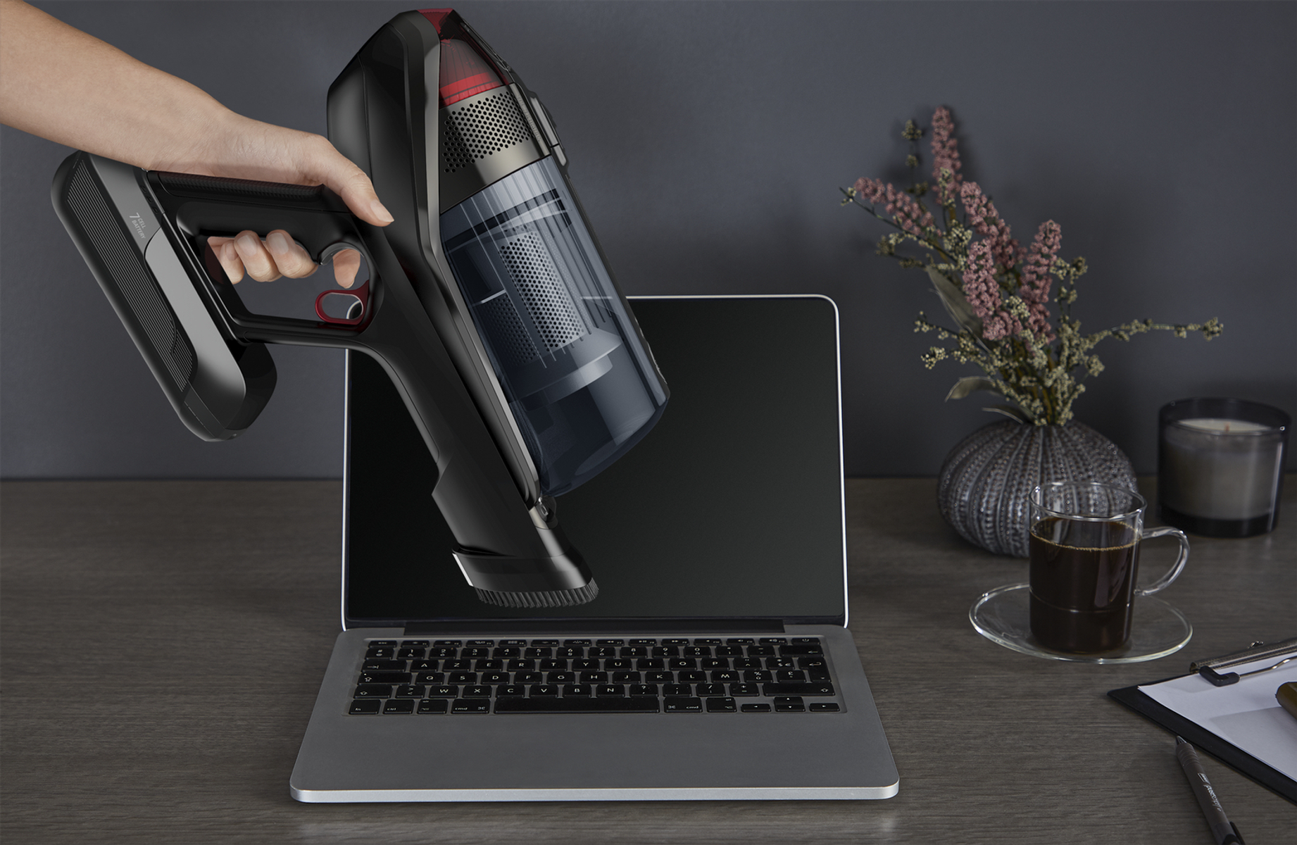 Mano sosteniendo una aspiradora negra sobre una computadora portátil. Escritorio con café, flores y bloc.