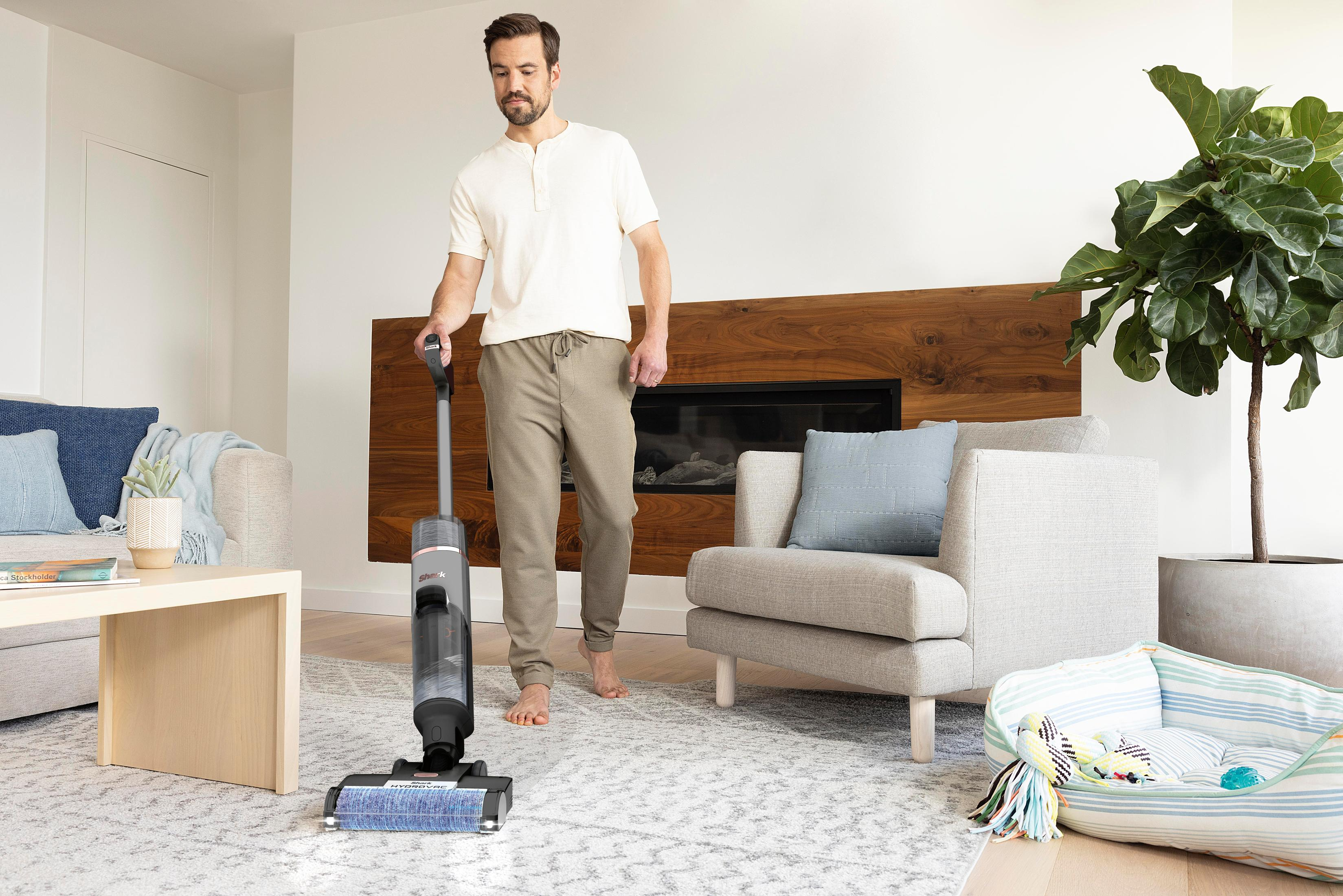 Un homme utilisant un aspirateur sans fil sur un tapis dans un salon avec un panier pour chien.