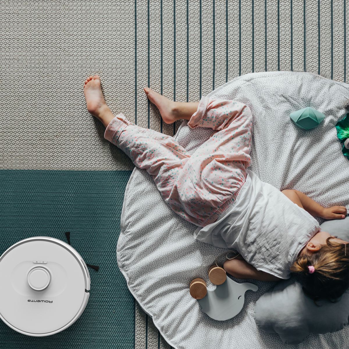 Dziecko leżące na białej macie z robotem sprzątającym w pobliżu na dywanie.