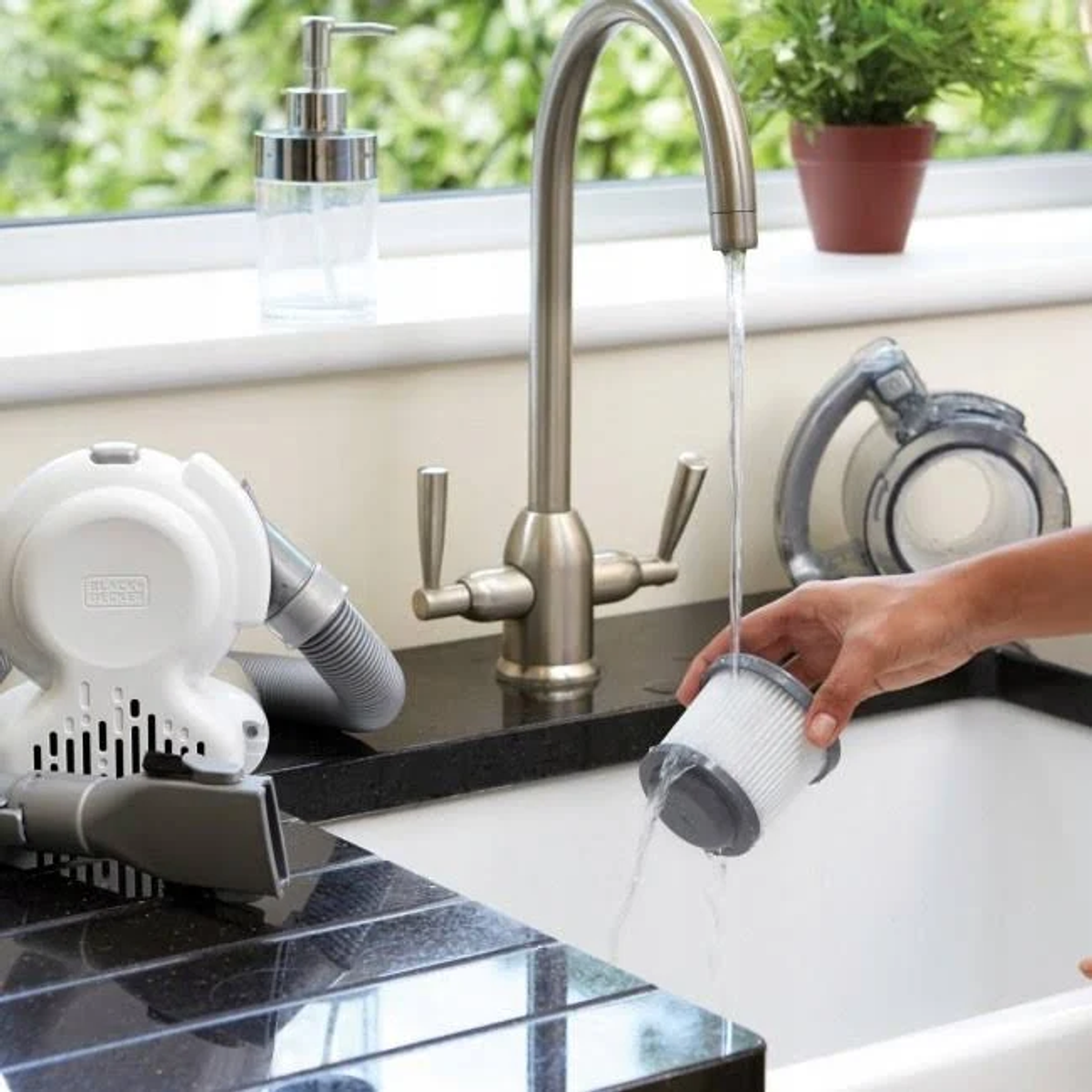 Une main rince un filtre sous un robinet. Un aspirateur blanc est à gauche.