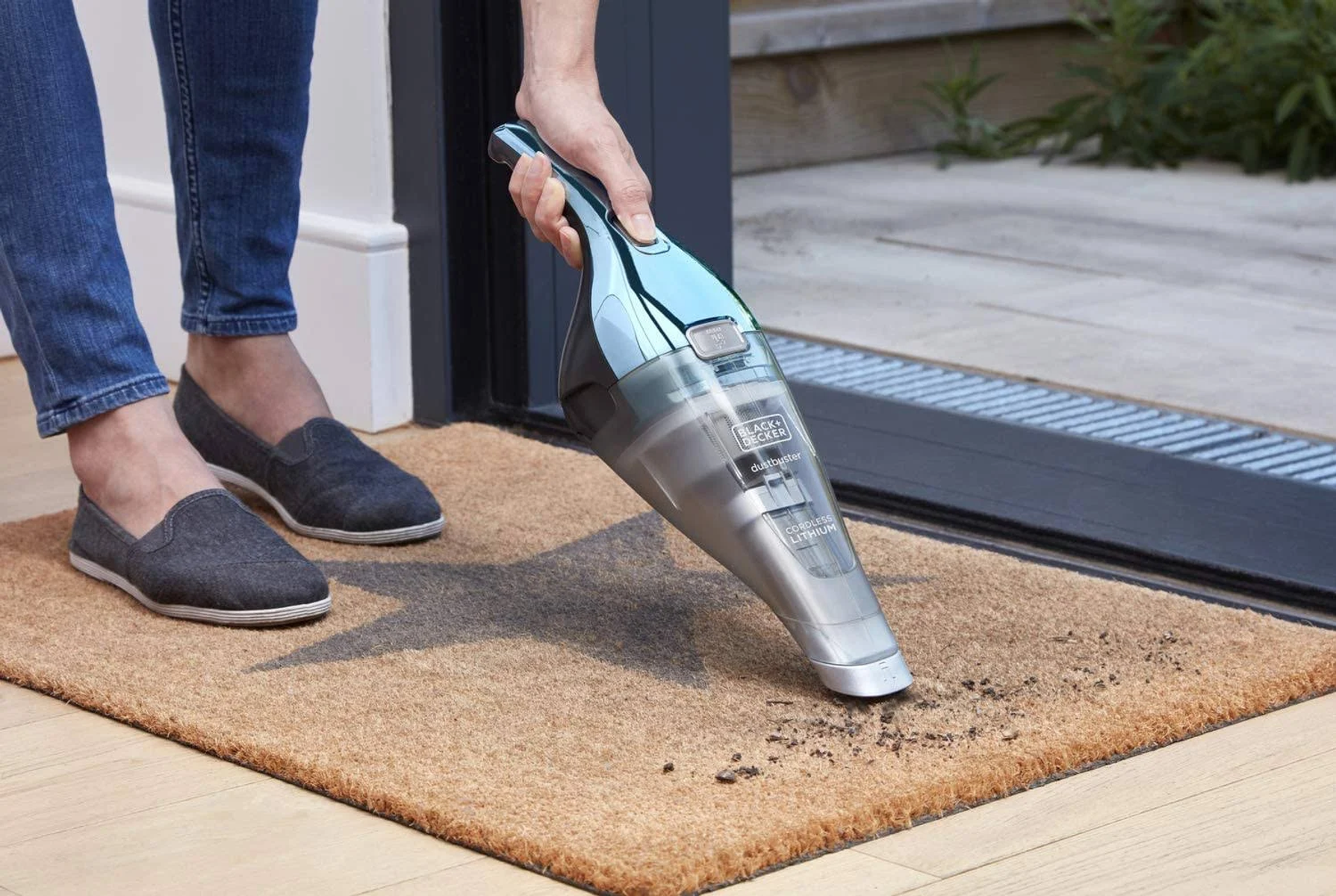 Osoba odkurza wycieraczkę odkurzaczem ręcznym, przed drzwiami.