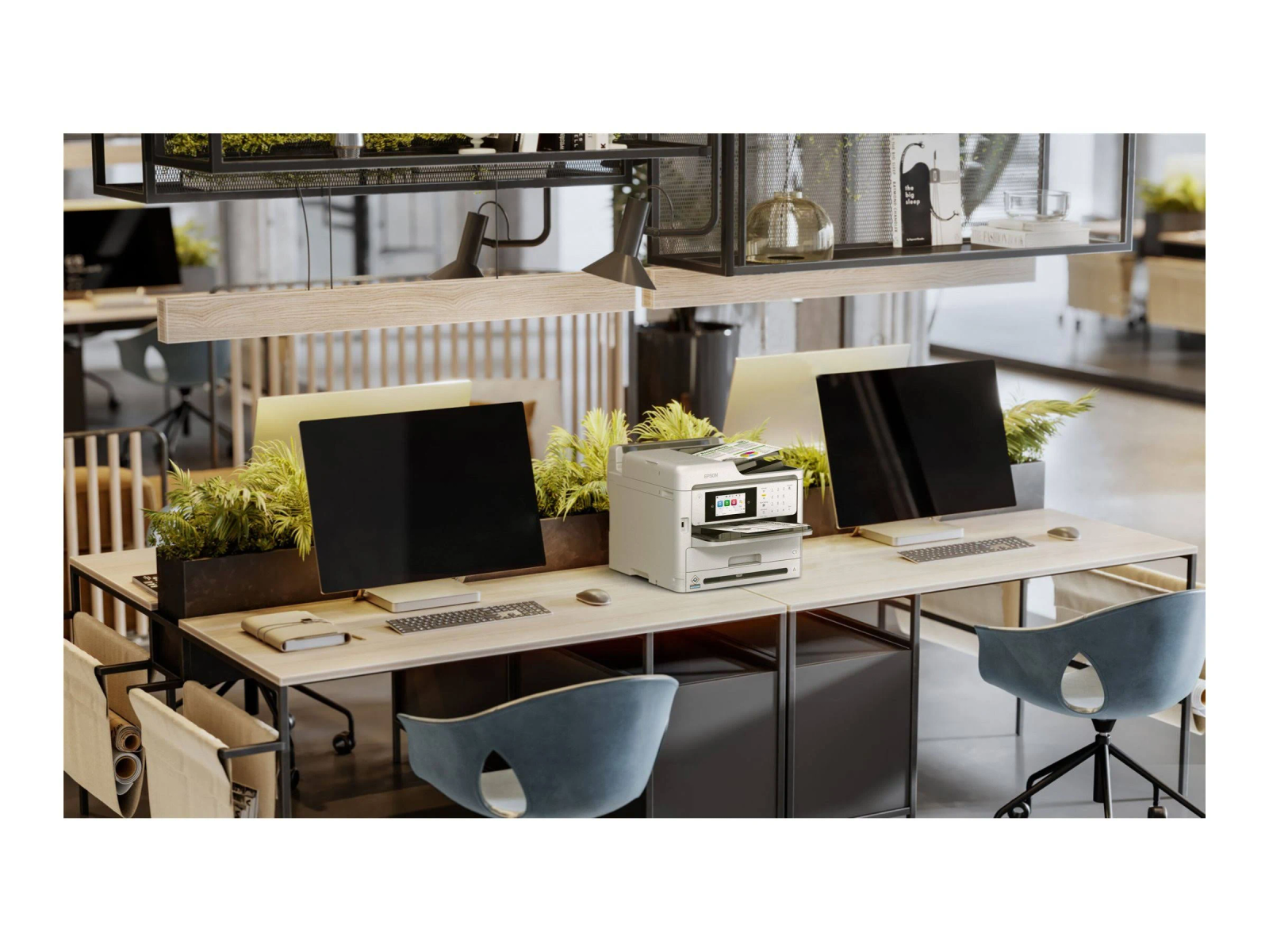 Przestrzeń robocza w biurze z komputerami, monitorami, drukarką i roślinami. Widoczne biurka i krzesła.