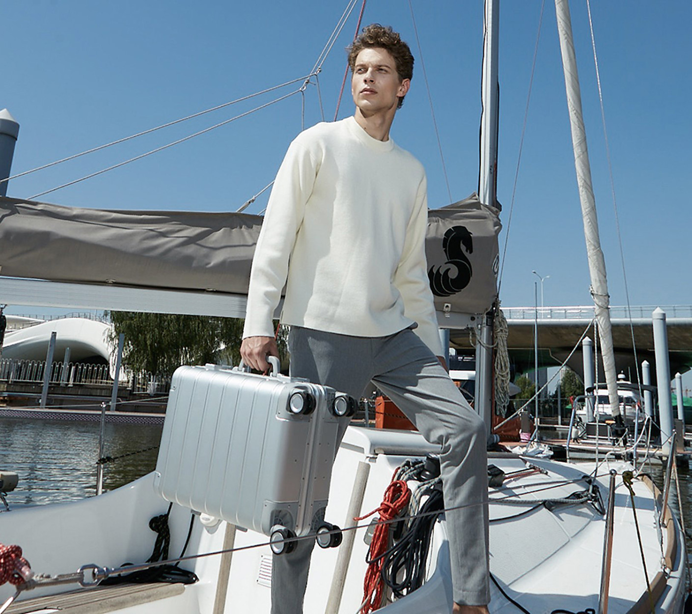 Ein Mann in weißem Pullover und grauer Hose hält einen silbernen Koffer auf einem Boot.