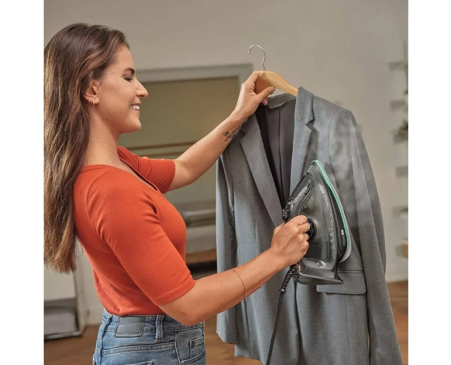 Frau bügelt einen grauen Blazer mit Dampf. Sie trägt ein orangefarbenes Hemd und Jeans. Hintergrund ist ein Raum.